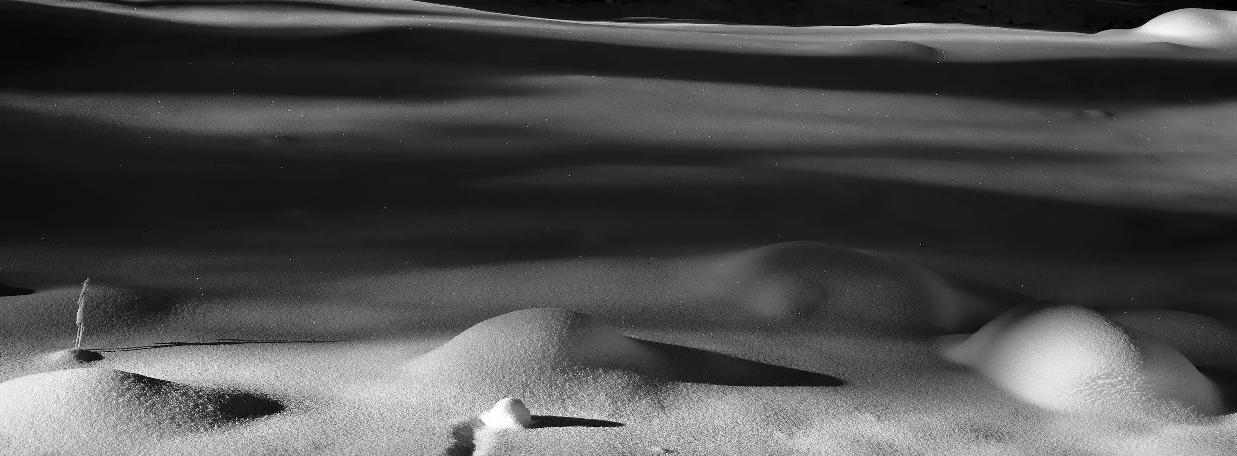 Black and white photo of snow-covered ground with smooth, rounded snow drifts and a faint line of rocky cliffs in the background.