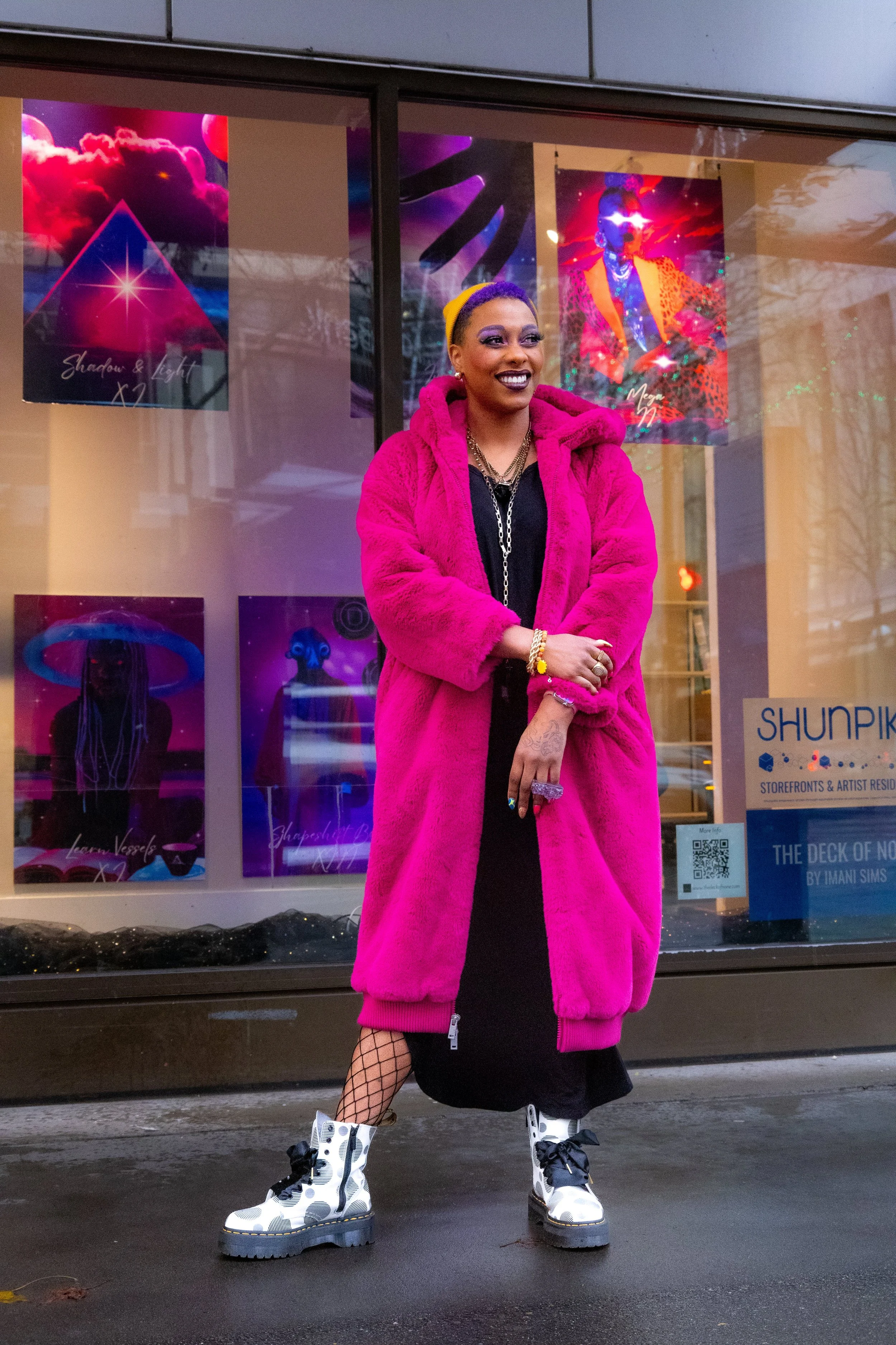 A woman standing outside in front of a window display with colorful artwork. She is wearing a bright pink fluffy coat, fishnet stockings, and white combat boots with black polka dots. She has dark makeup, jewelry, and a yellow headband, smiling with arms crossed.
