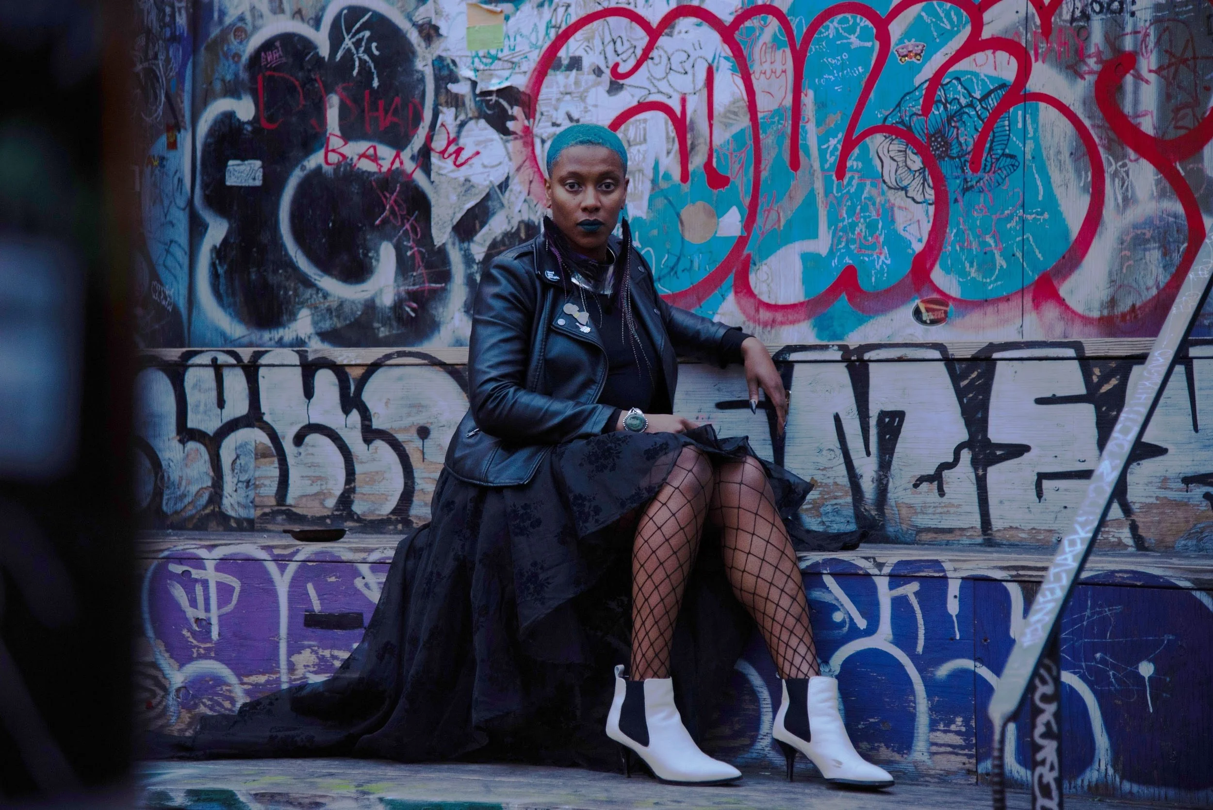 Imani sitting in front of a grafitti wall wearing a black skirt and black leather jacket