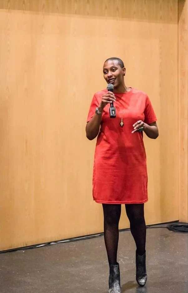 A woman with short hair standing on a stage, holding a microphone, wearing a red dress, fishnet tights, and black boots.