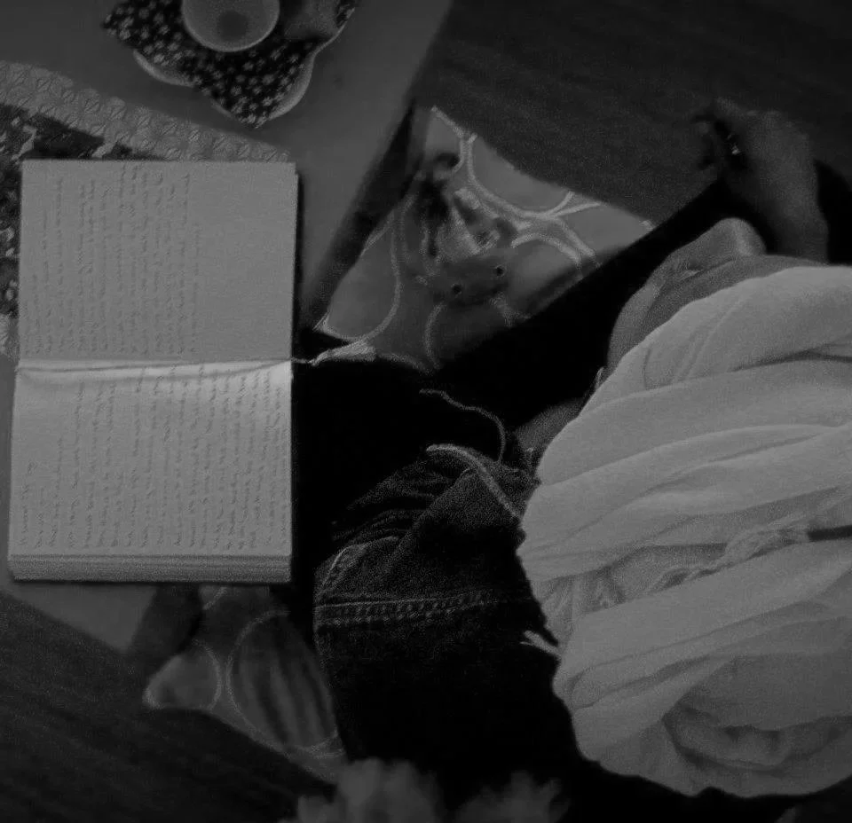 A person sitting on the floor reading a book, with a cup nearby and some headphones resting on the floor. The person is wearing a white shirt and denim shorts.