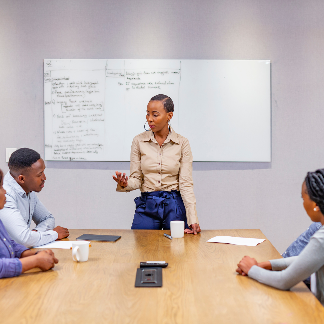 A woman standing at a conference table giving a presentation to four seated individuals in a meeting room with a whiteboard behind her.