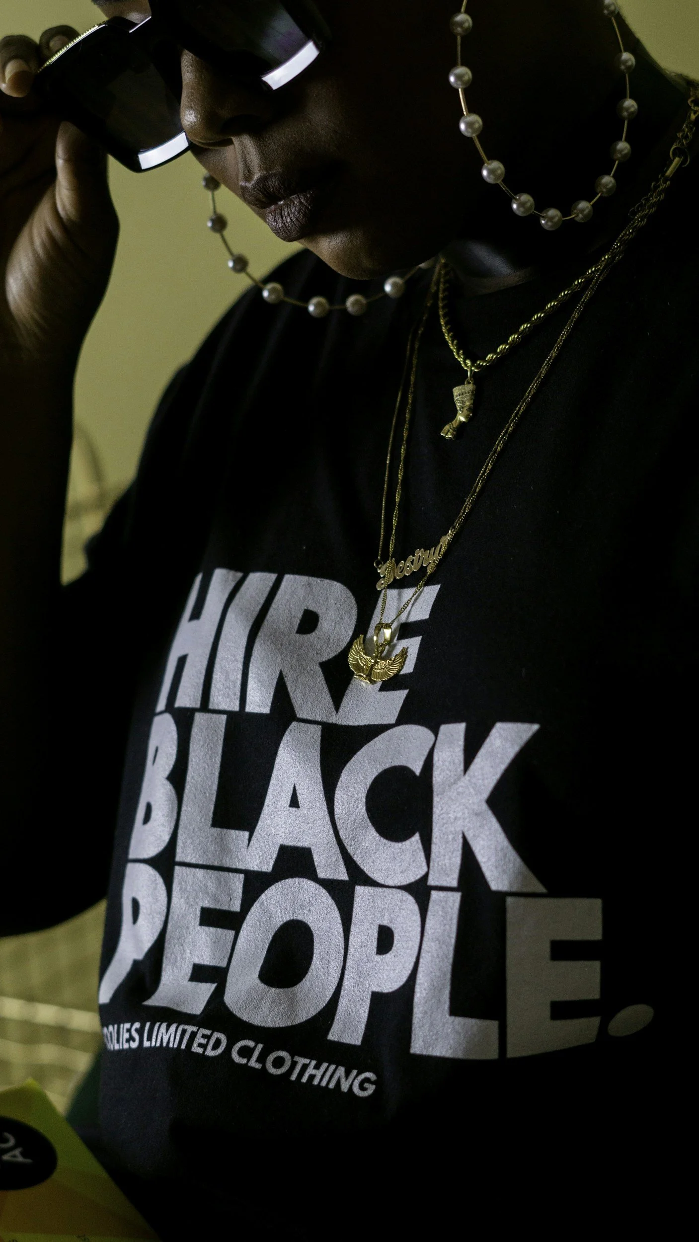 Person wearing a black t-shirt with white bold text stating "HIRE BLACK PEOPLE." The person has layered gold necklaces, one with a pendant of a winged figure, another with a nameplate. They are holding a camera or binoculars close to their face, wearing black sunglasses with reflective lenses. They also have pearl earrings and a pearl necklace.