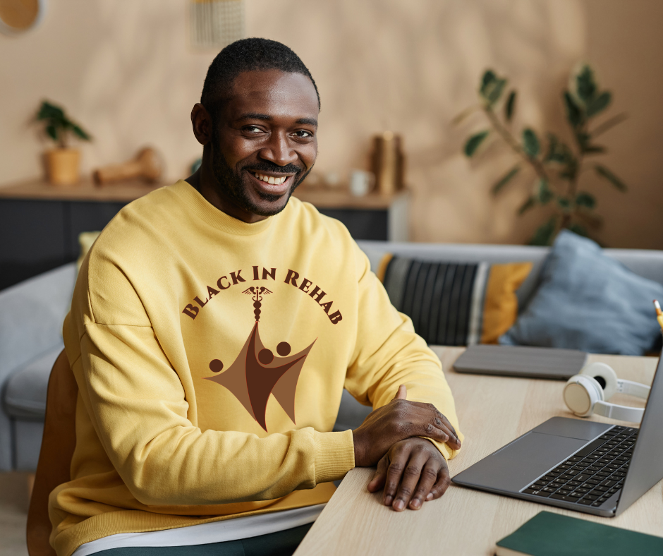 A smiling man wearing a yellow sweatshirt with the text 'Black In Rehab' and a graphic of three abstract figures. He sits at a desk with a laptop, headphones, and a notepad, in a cozy room with plants and decorative pillows.