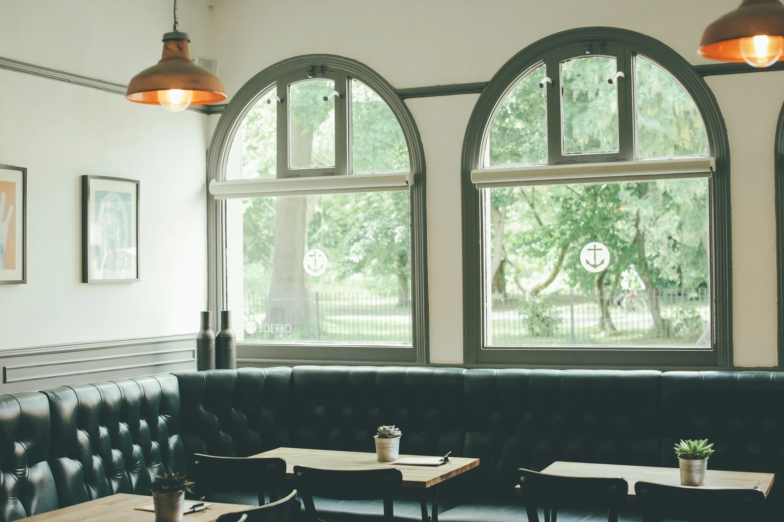 Interior of a cozy café with large arched windows, green trim, and greenery outside. Black leather seating, wooden tables with small potted succulents, and pendant lights hanging from the ceiling.