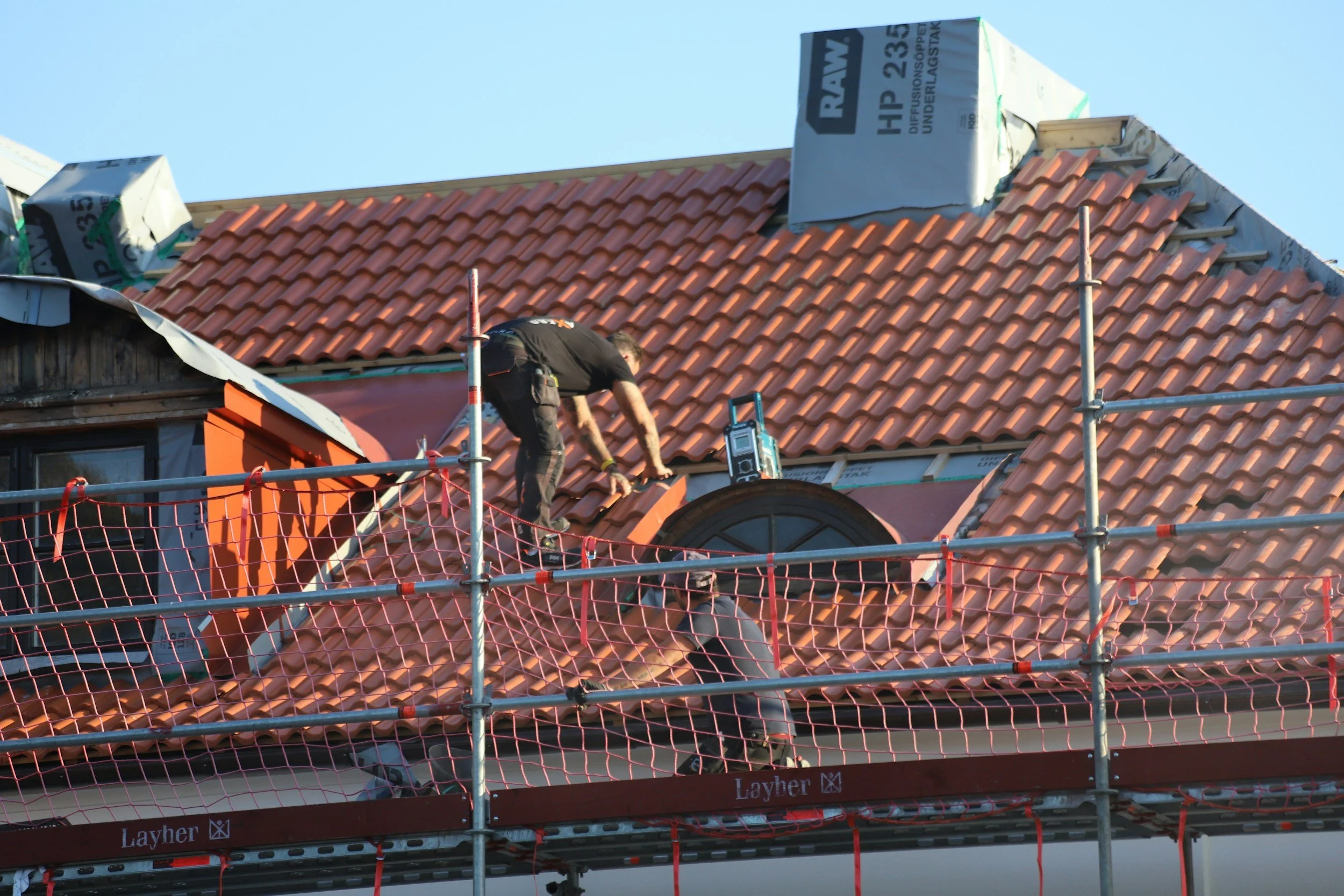 Two workers installing red roof tiles on a building under construction, with scaffolding around the roof.