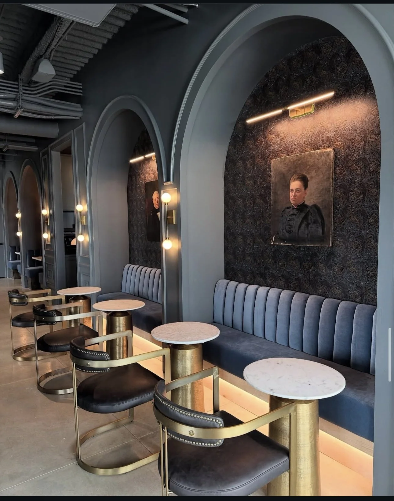 Elegant interior of a cafe or lounge with gray upholstered benches, black and gold chairs, and marble-topped tables. Portraits hang on dark textured walls, illuminated by modern lighting fixtures.