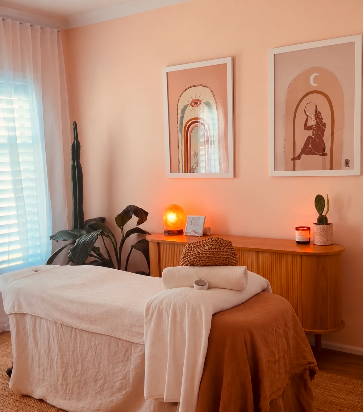 A cozy massage therapy room with a massage table draped in white linen. There is a rolled towel on the end of the table, and a woven pillow. On the wooden side table, there is a salt lamp, a framed picture, a potted cactus, and a candle. The wall behind features two framed art prints, one with an eye and rainbow design, and the other with a woman sitting with a crescent moon. Large plants are near a window with sheer curtains, and the room has warm, ambient lighting.