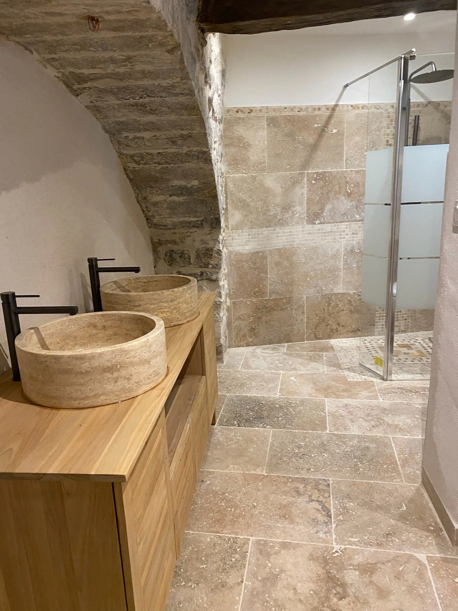 Bathroom with rustic stone and tile design, featuring a wooden vanity with two round stone vessel sinks, and a glass shower with a frosted glass panel, stone-tiled walls, and a pebble mosaic shower floor.