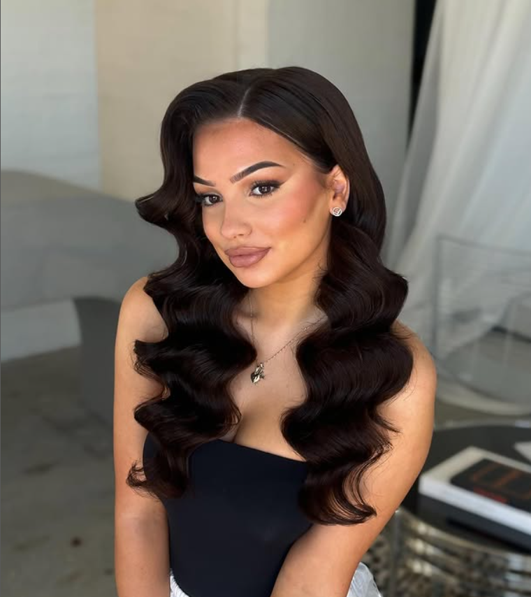 A woman with long, dark, wavy hair, makeup, earrings, and a necklace, posing indoors.