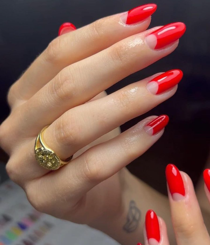 Close-up of a hand with red and nude ombré nails, wearing a gold ring, with a tattoo of a rose on the wrist.