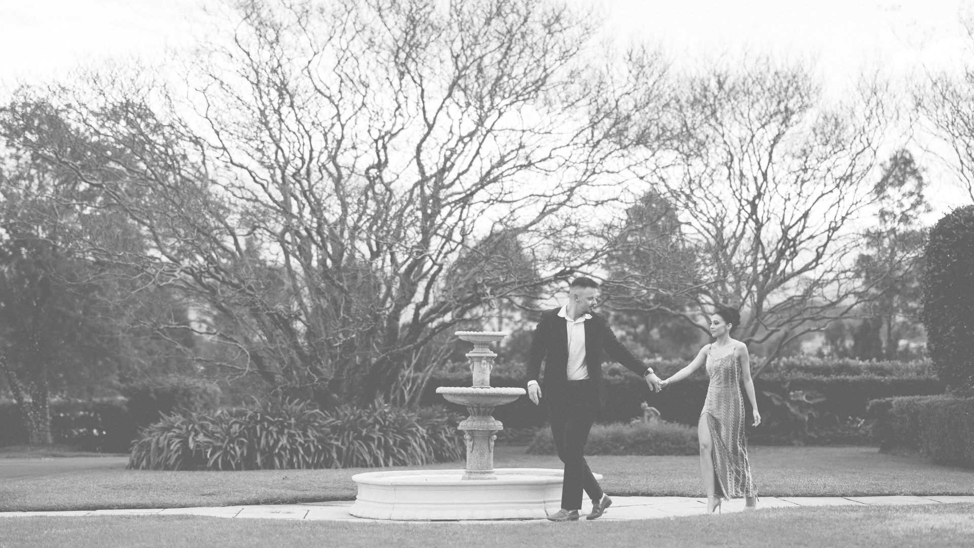 A black and white photo of a man and woman holding hands near a fountain in a park with trees in the background.