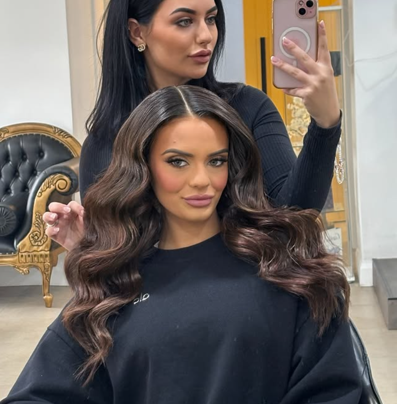 A woman with long, wavy brunette hair and makeup sits in a salon chair, taking a selfie in a mirror. A stylist with black hair, wearing a black top, is behind her, holding a phone to photograph the client. The salon has a vintage-style black and gold chair in the background.