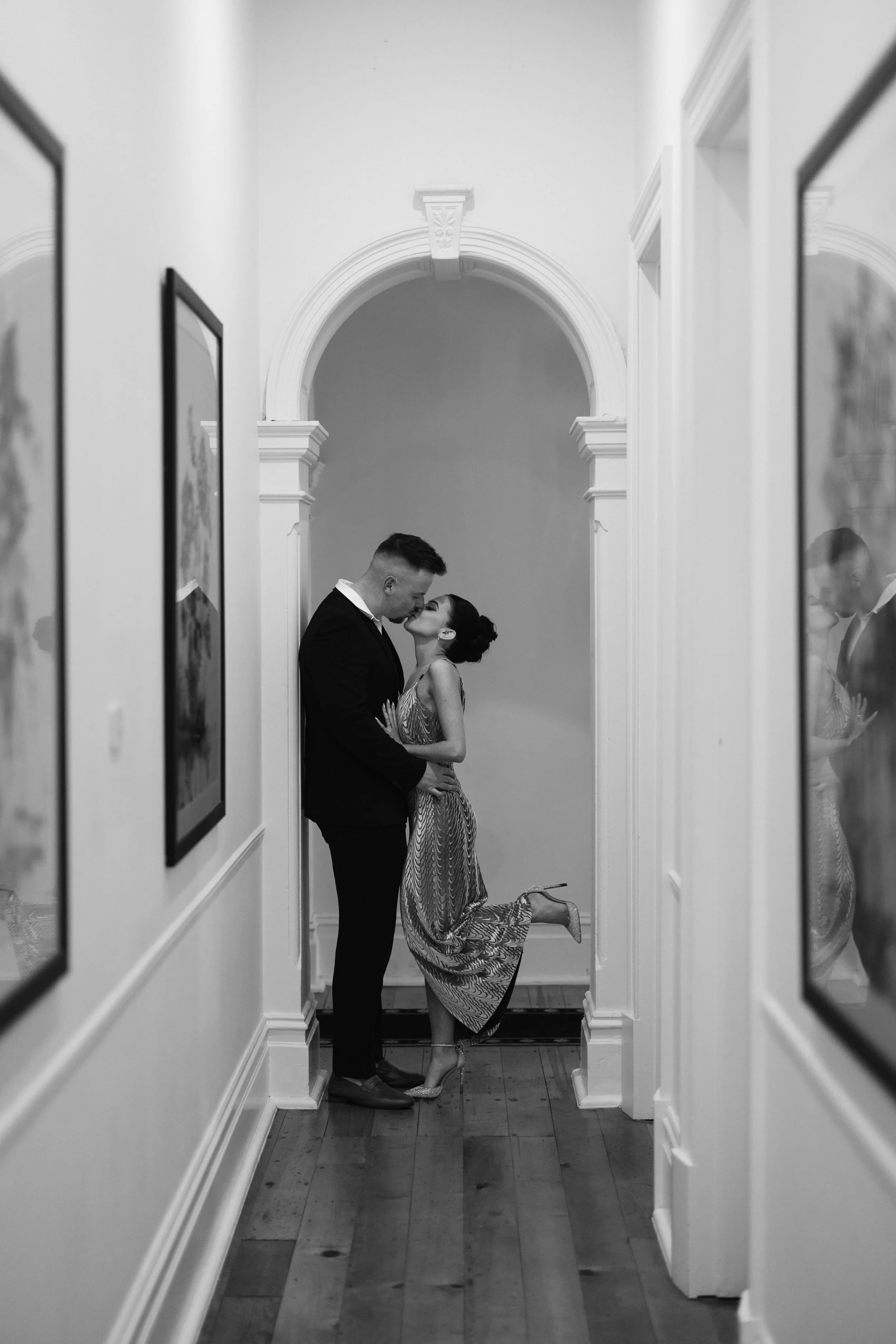 A black and white photograph of a man and woman embracing in a narrow hallway with framed artwork on the walls. The woman is lifting her right leg and leaning into the man as they share an intimate moment.