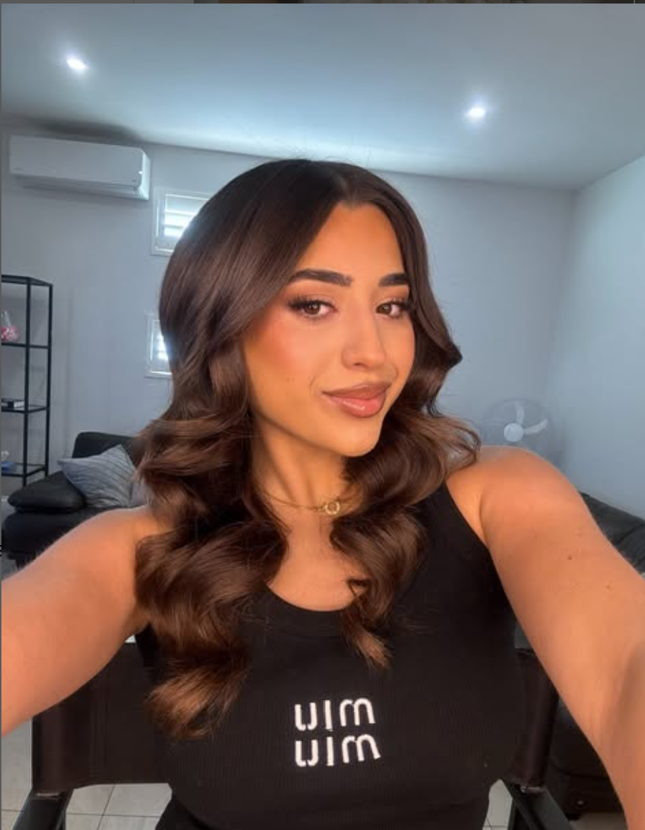 A woman with long, wavy brown hair taking a selfie in a modern living room with white walls, a black fabric chair, a gray throw blanket, and windows with blinds.