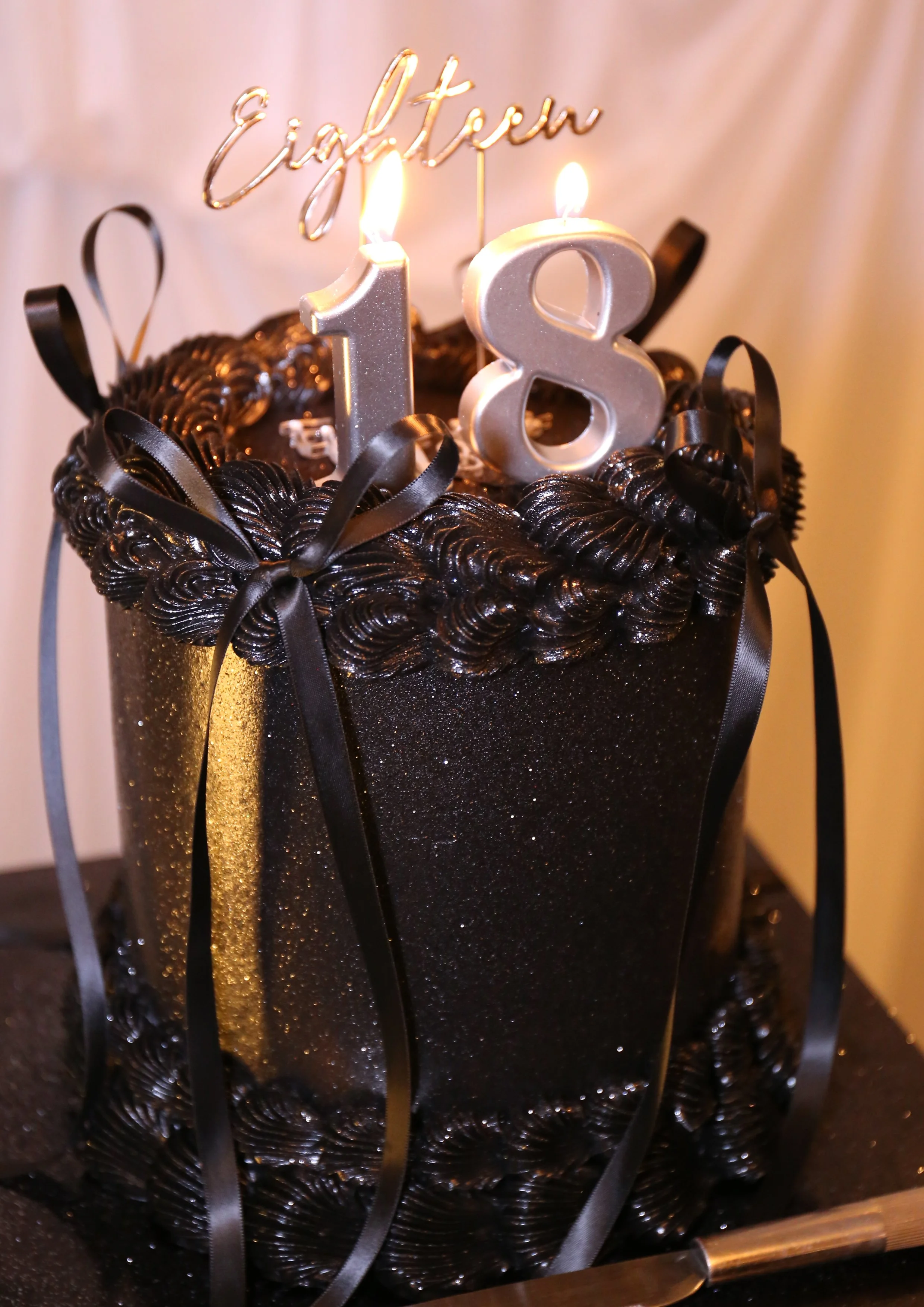 Black birthday cake with black piping and ribbons, gold stripe on the side, topped with silver "18" candles and a "Happy Eighteen" cake topper with lit candles.