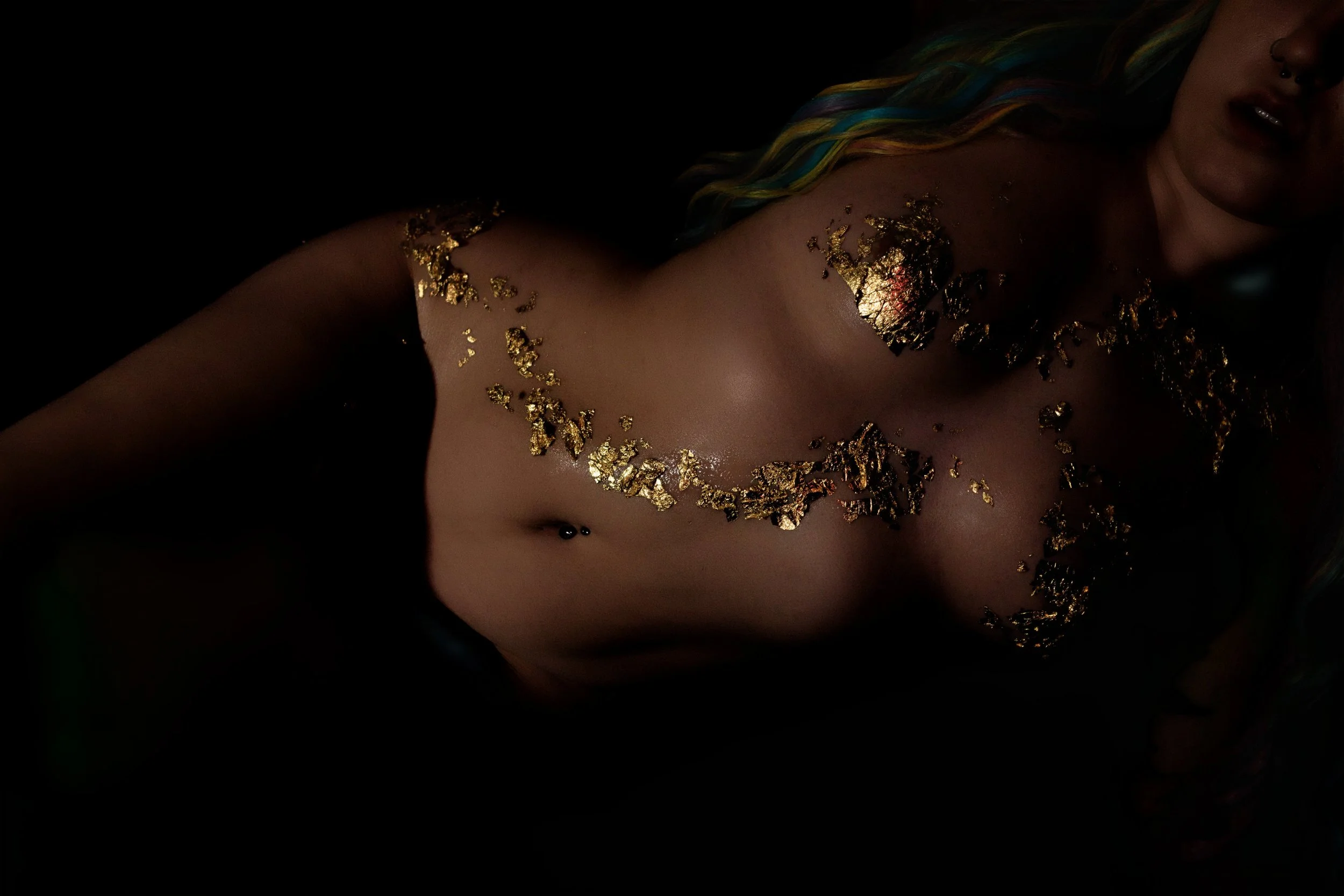 A topless woman with gold leaf applied to her upper chest and shoulders, partially visible with hair containing colorful streaks and a dark background.