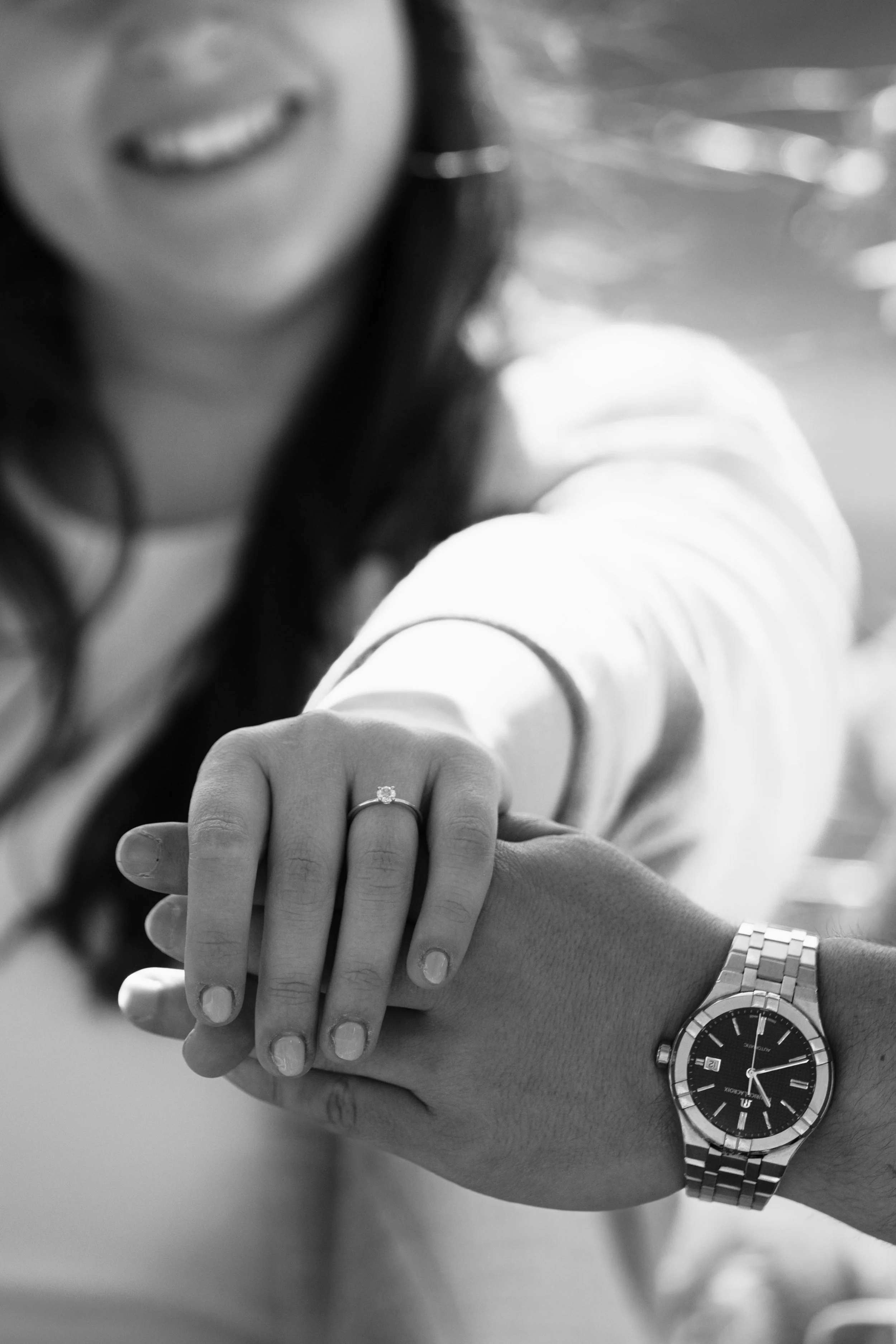 Ein Portrait einer lächelnden Frau, die ein Paar Hände hält, wobei eine Frau ein Verlobungsring zeigt und eine Person eine Armbanduhr trägt.