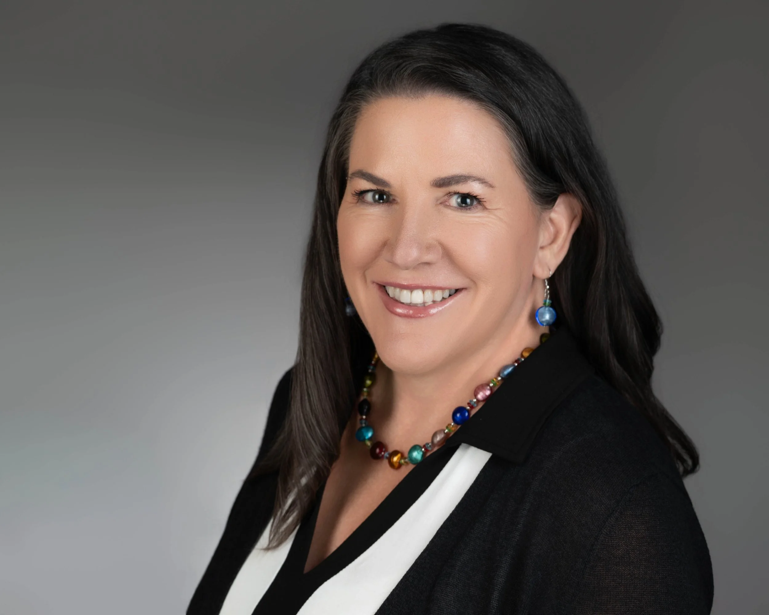 A woman with dark hair smiling, wearing a black and white top, colorful necklace, and matching earrings, against a gray background.