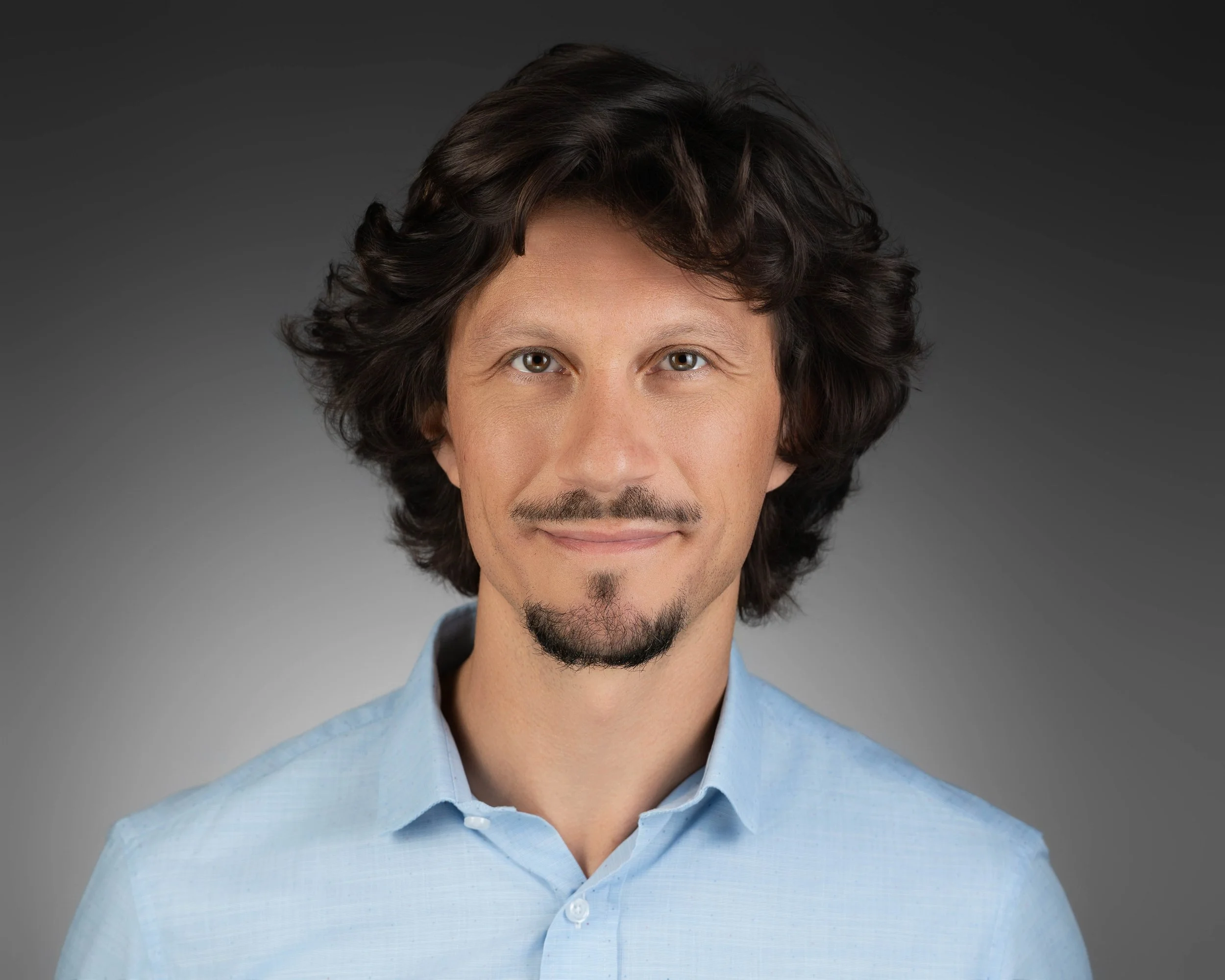 Portrait of a man with wavy dark hair, light skin, and a slight smile, wearing a light blue shirt with a gray gradient background.
