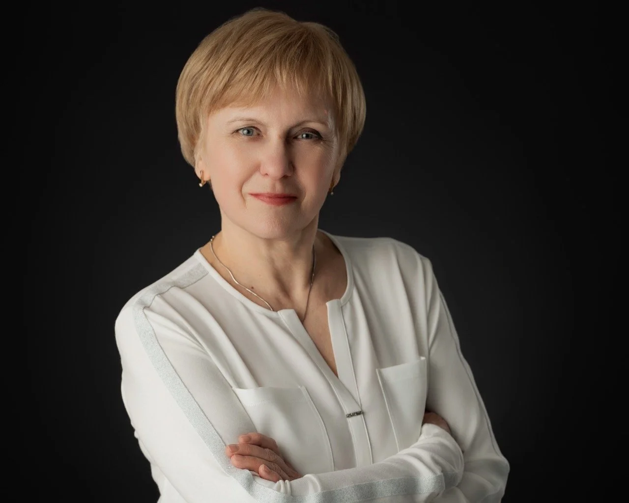 A woman with short blonde hair, wearing a white blouse with silver accents, standing with arms crossed against a dark background.