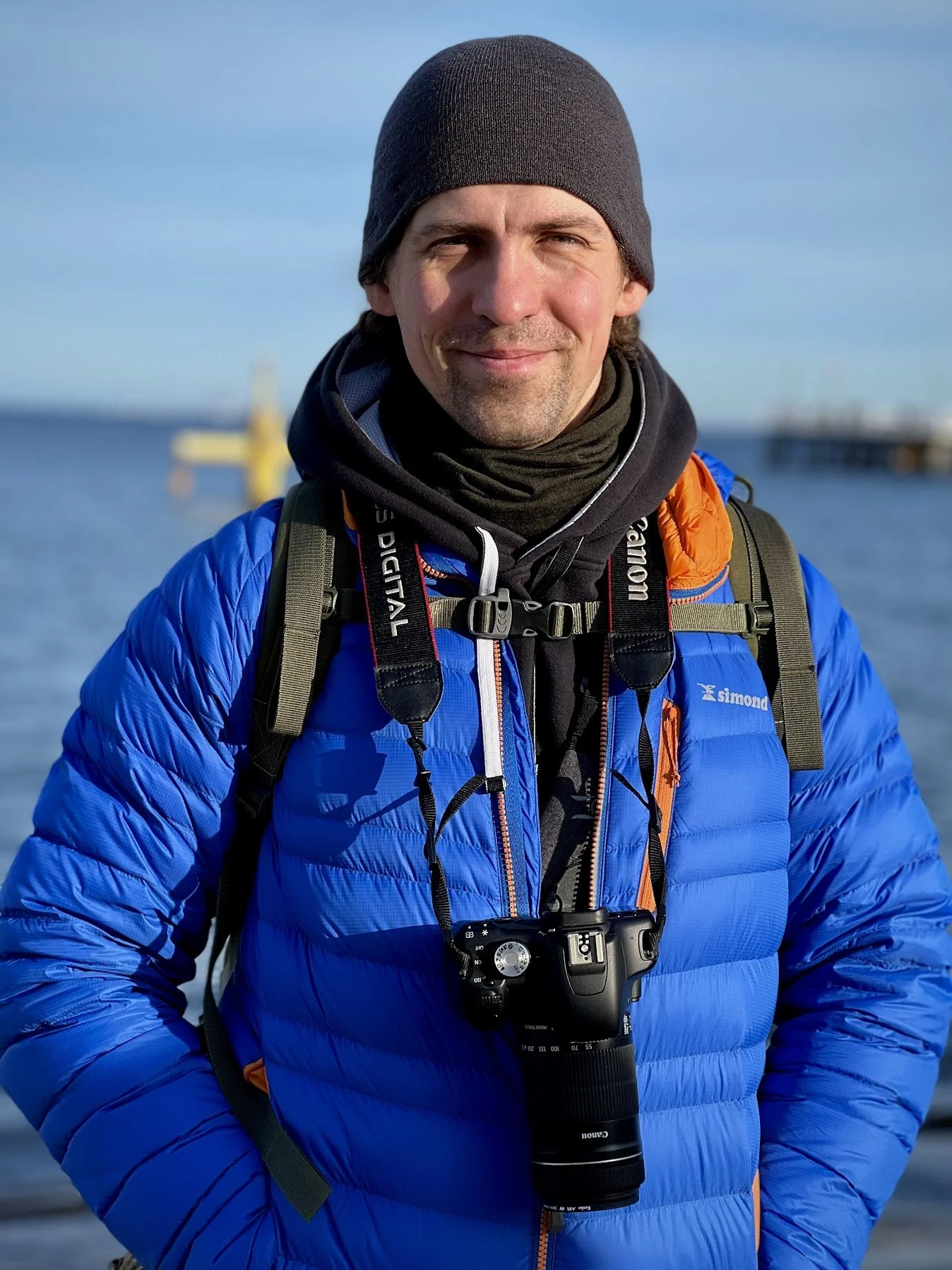 Outdoor portrait of a man wearing a blue jacket and beanie with a camera around his neck by the water