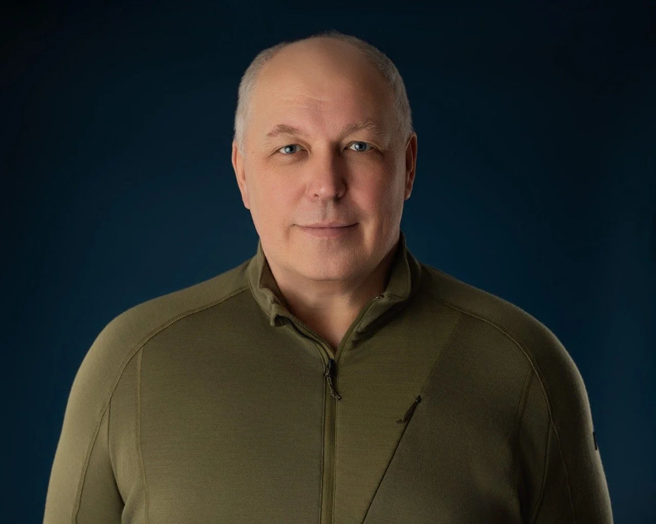 Professional headshot of a man in an olive zip sweater on a blue background