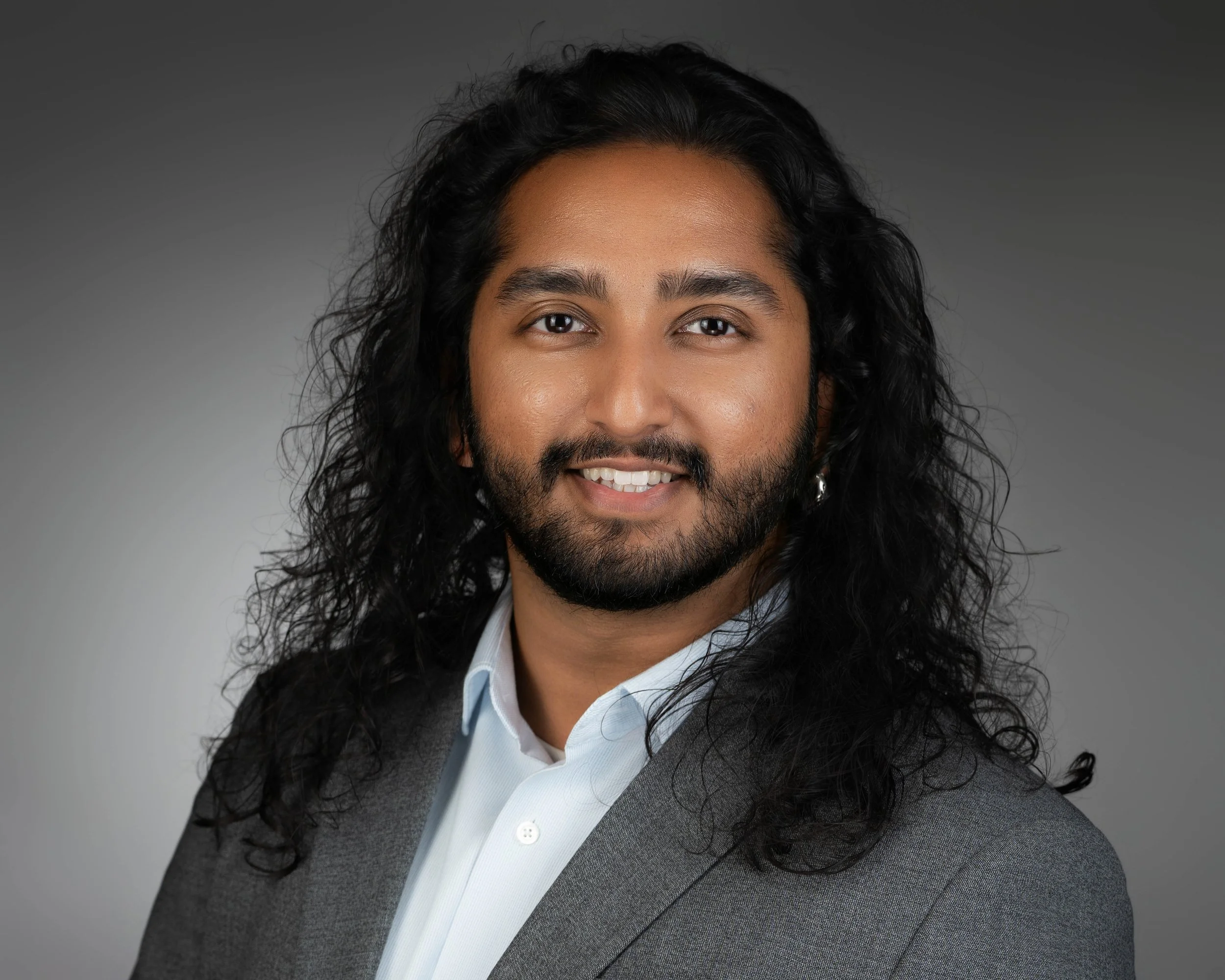 Professional portrait of a man with long curly black hair, a beard, wearing a light blue collared shirt and gray suit jacket, smiling against a gray gradient background.