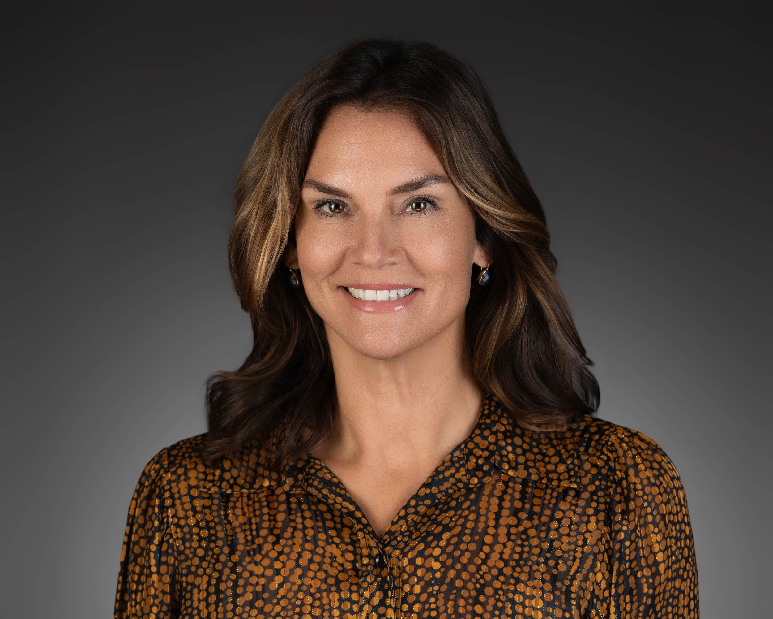 Portrait of a woman with shoulder-length brown hair, wearing a patterned blouse and earrings, smiling against a gray gradient background.
