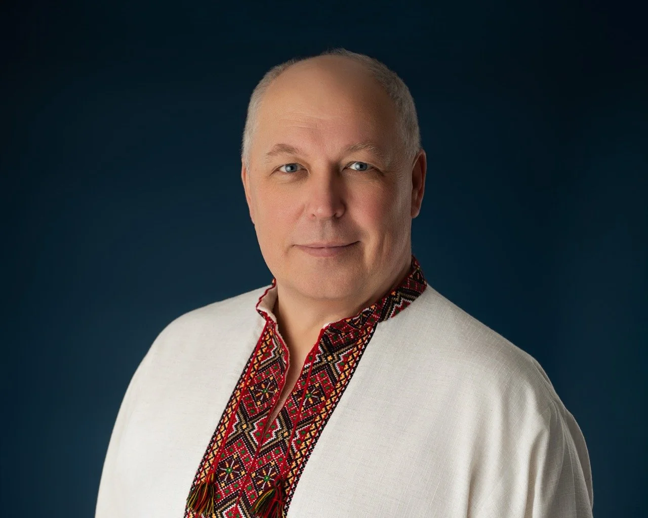 Portrait of a middle-aged man with a friendly smile, wearing traditional embroidered clothing, against a dark background.