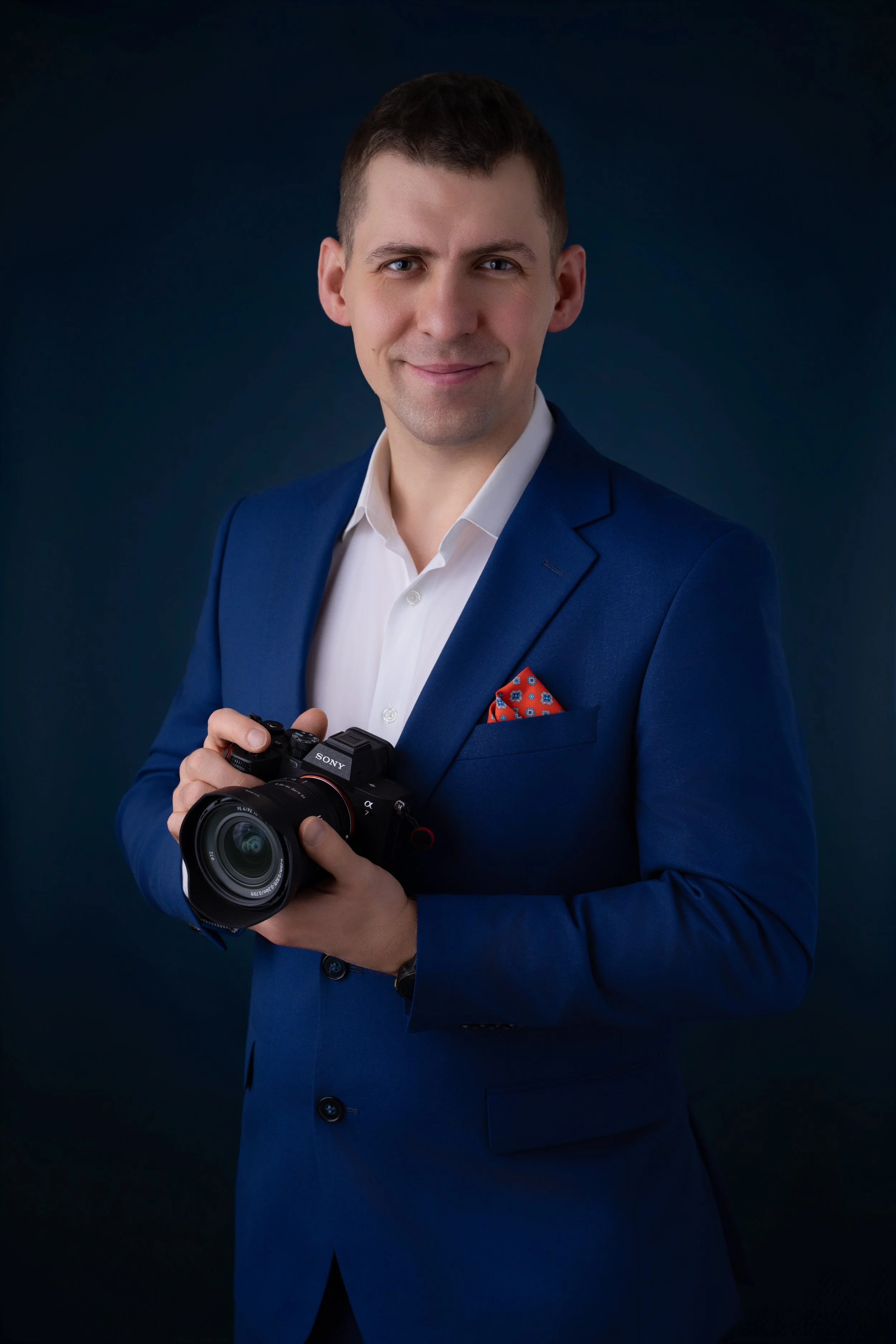 A man in a blue suit holding a camera, smiling, against a dark background.