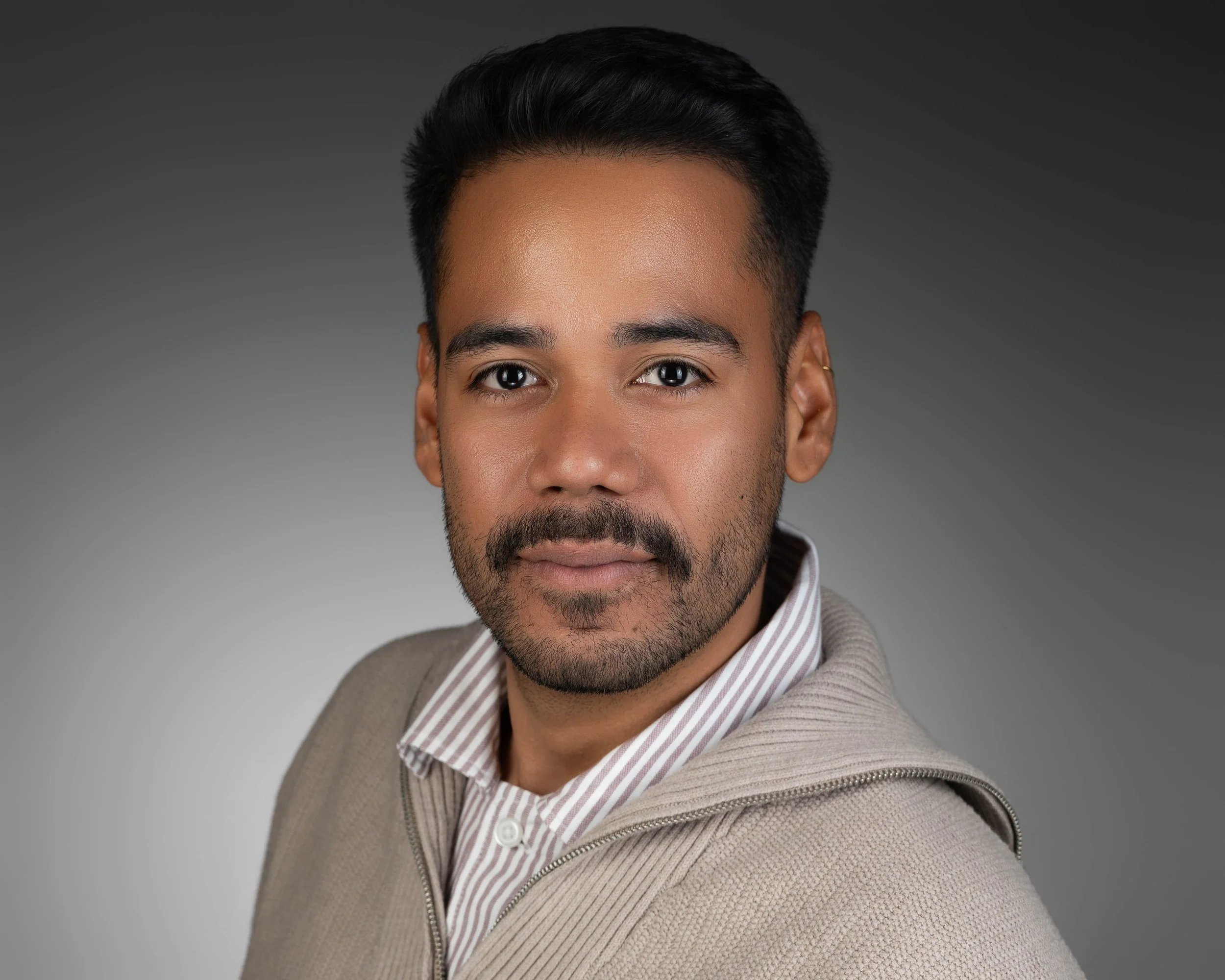 Close-up portrait of a man with short black hair, a beard, wearing a striped shirt and beige sweater, against a gradient gray background.