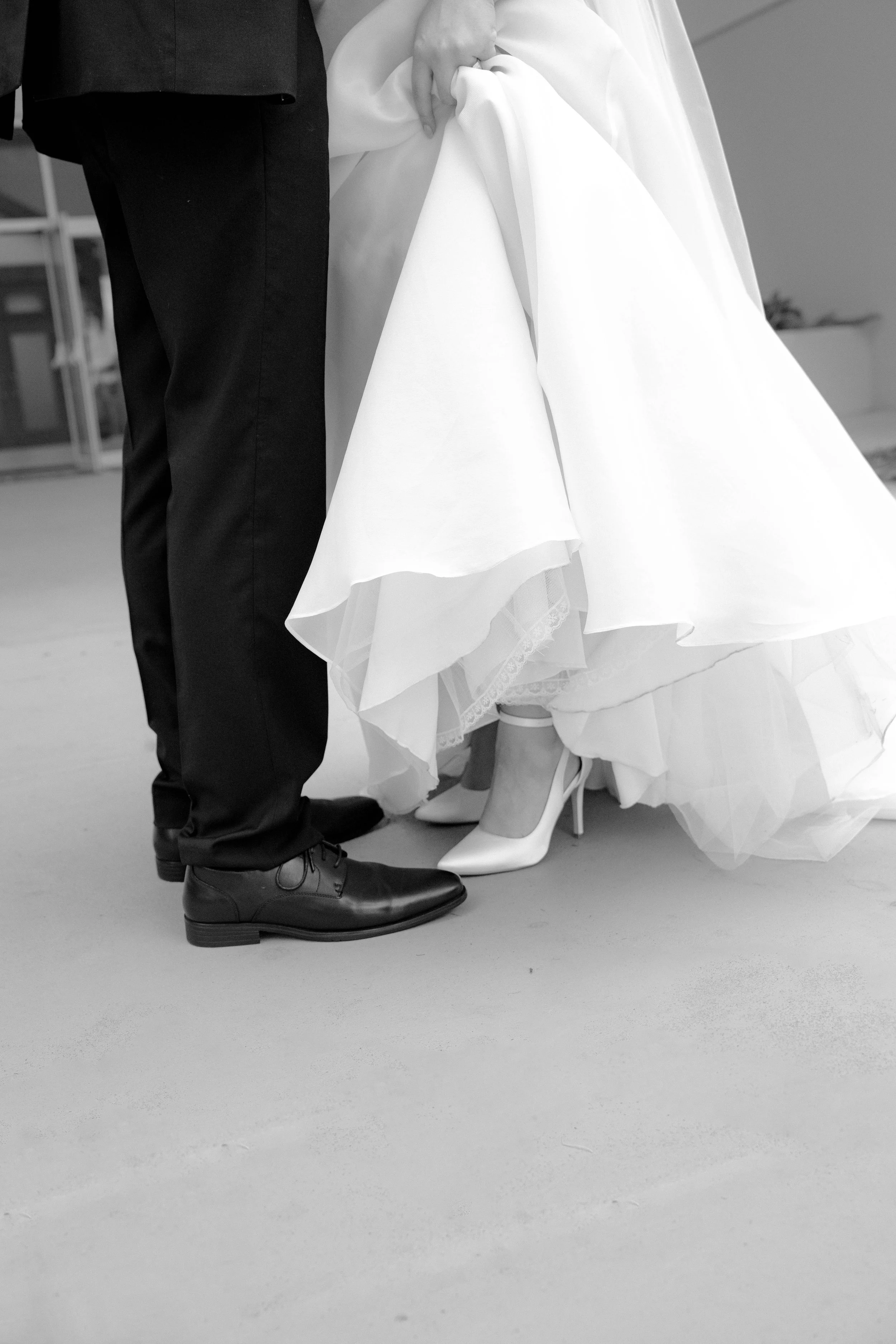 Close-up of a couple's legs and feet, the woman in a wedding dress and heels, the man in black dress shoes and dark suit, holding hands.