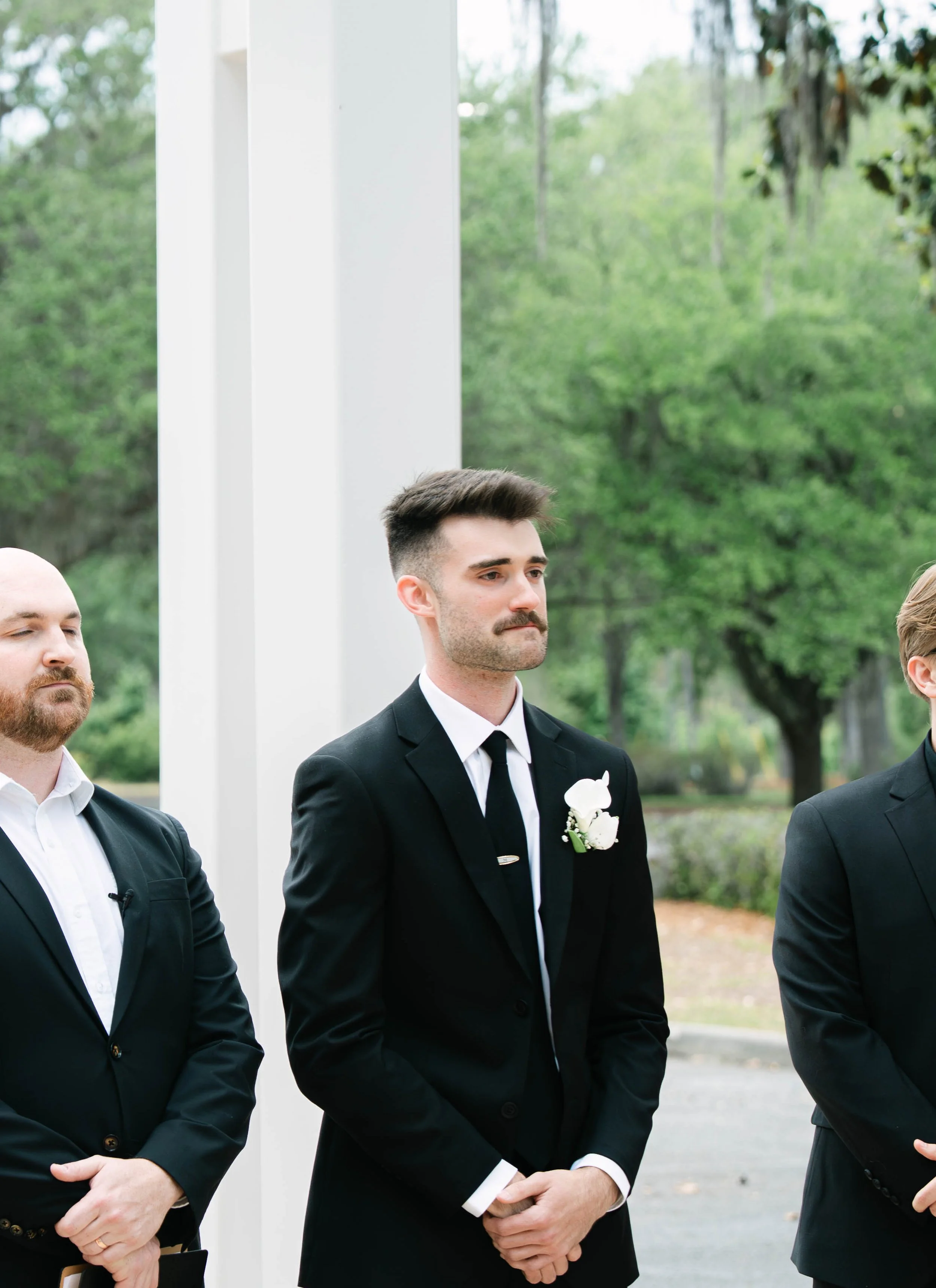 Three men standing outdoors dressed in formal black suits, white shirts, and black ties, during a daytime event with trees in the background.