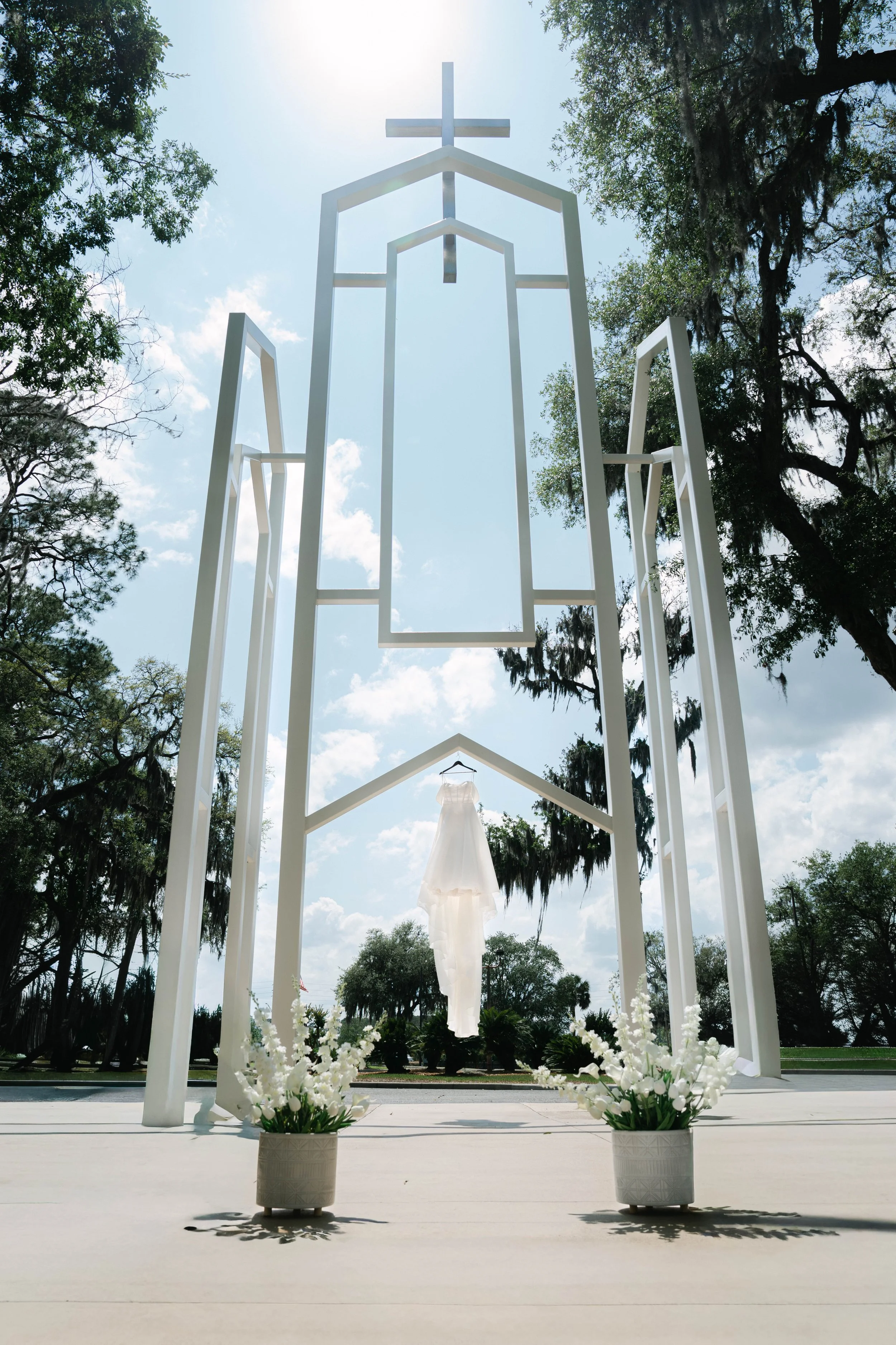 Outdoor wedding altar with white framework, hanging white dress, and white floral arrangements on the ground, set against a backdrop of trees and blue sky.