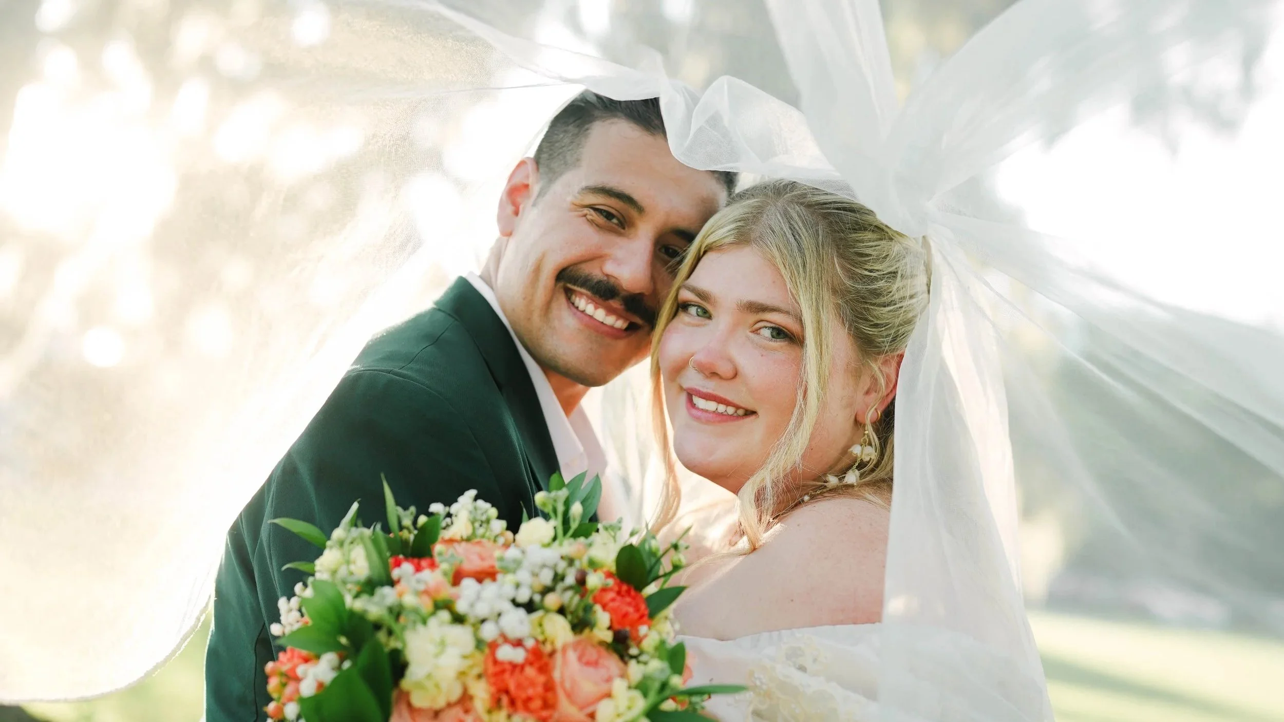 A smiling couple, a man in a dark suit and a woman in a white dress, close together with their heads touching under a veil, holding a bouquet of flowers, outdoors in a sunlit setting.