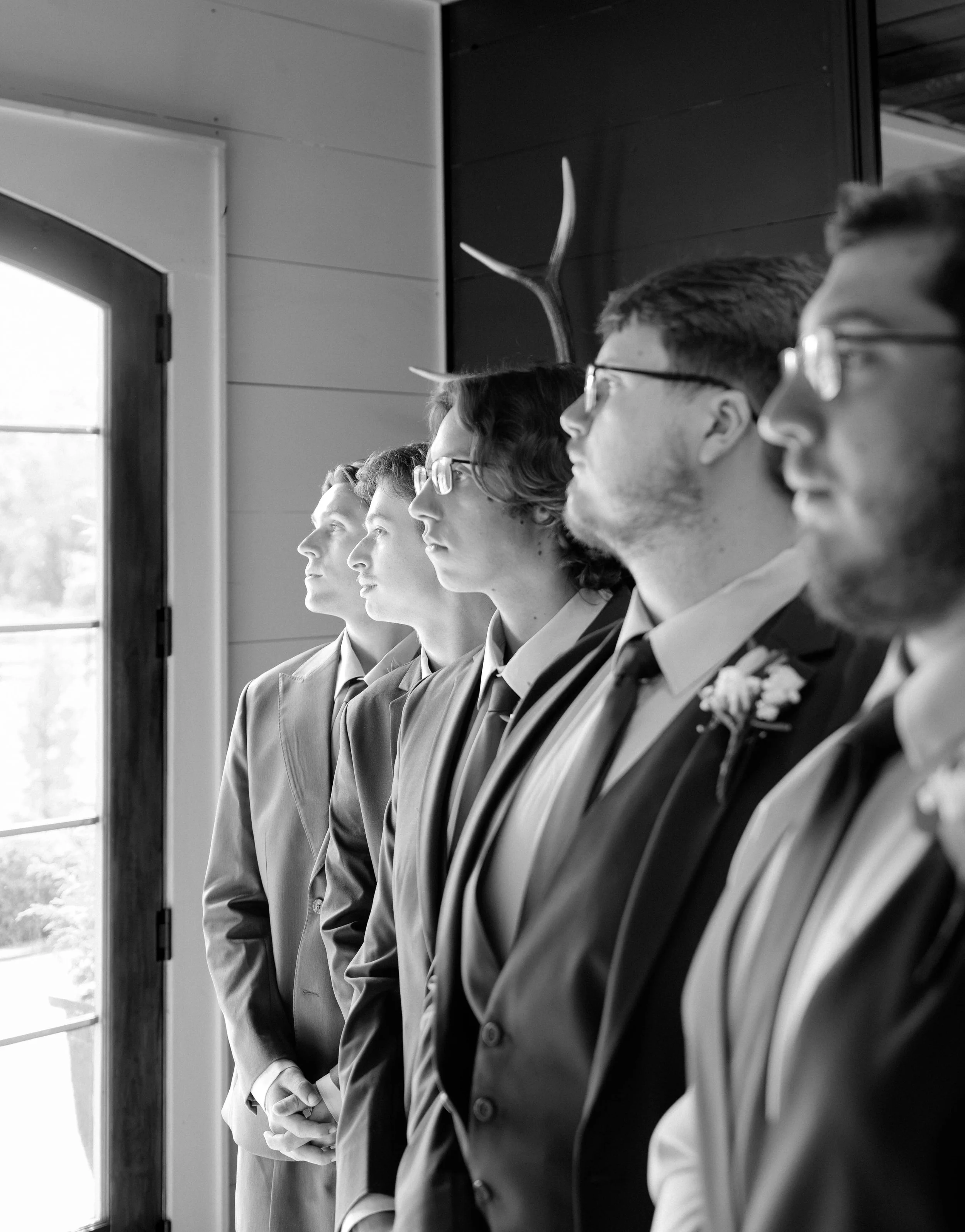 Line of people dressed in suits standing side by side near a window, facing right, in black and white.