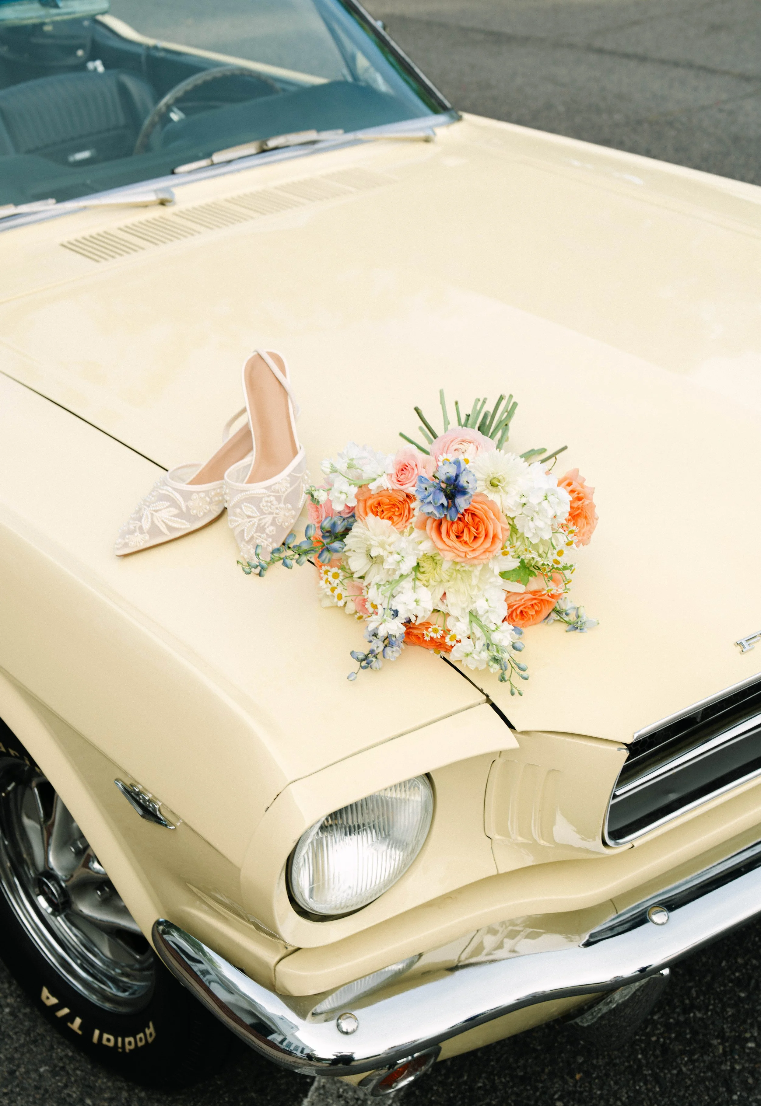 A vintage cream-colored car decorated with a bouquet of flowers and white high-heeled shoes on the hood.