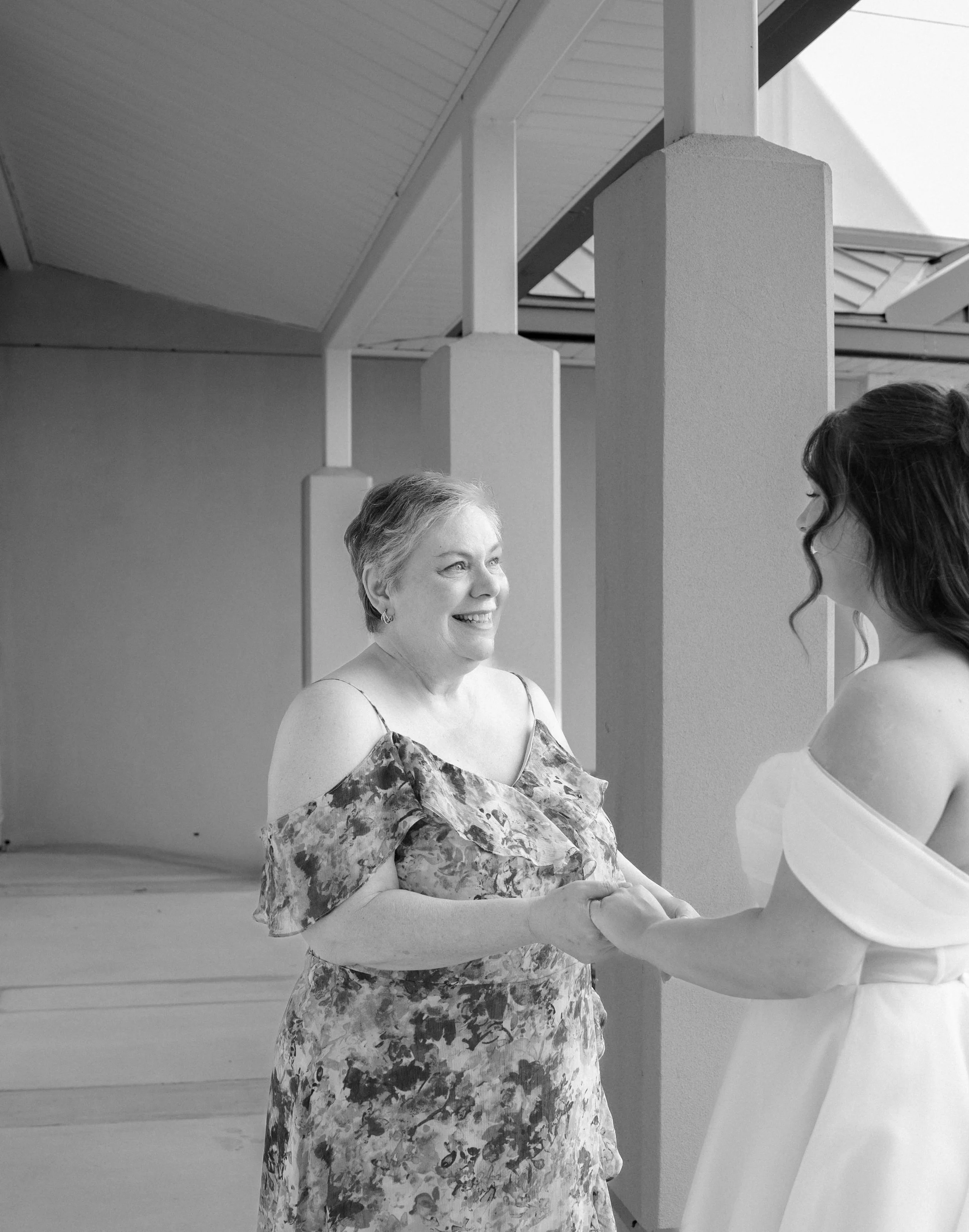 A middle-aged woman in a floral dress smiling and holding hands with a young woman in a light-colored dress, both facing each other indoors under a ceiling with beams and columns.