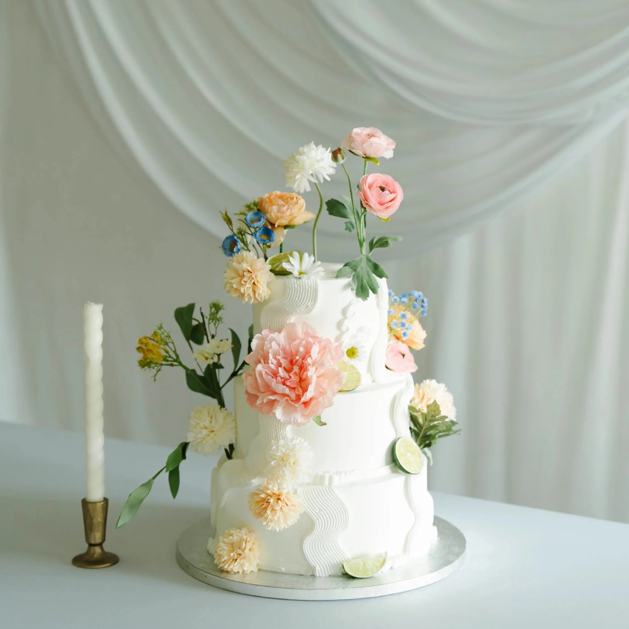 A white tiered wedding cake decorated with various pastel-colored flowers, lime slices, and greenery, with a backdrop of draped light-colored fabric.