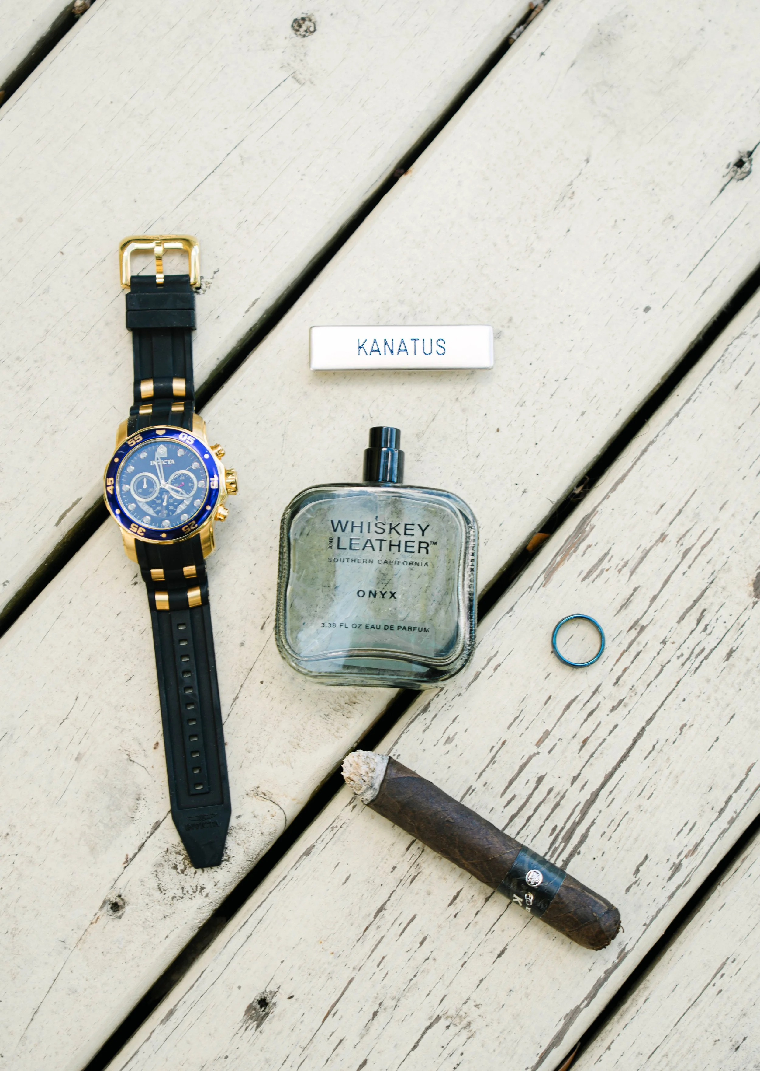 A black and gold wristwatch, a bottle of whiskey leather perfume, a rectangular white box labeled 'KANATUS,' a blue ring, and a cigar on a white wooden surface.
