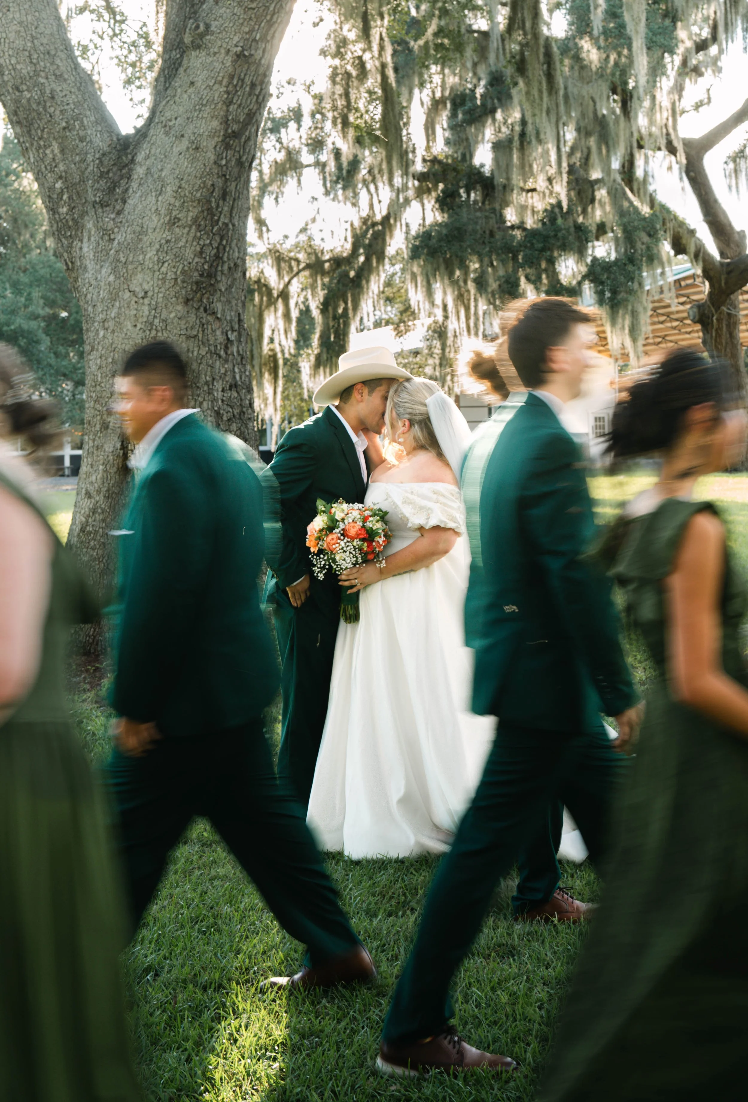 A bride and groom sharing a kiss outdoors during their wedding, surrounded by wedding guests walking by, with a large tree and moss hanging from its branches in the background.