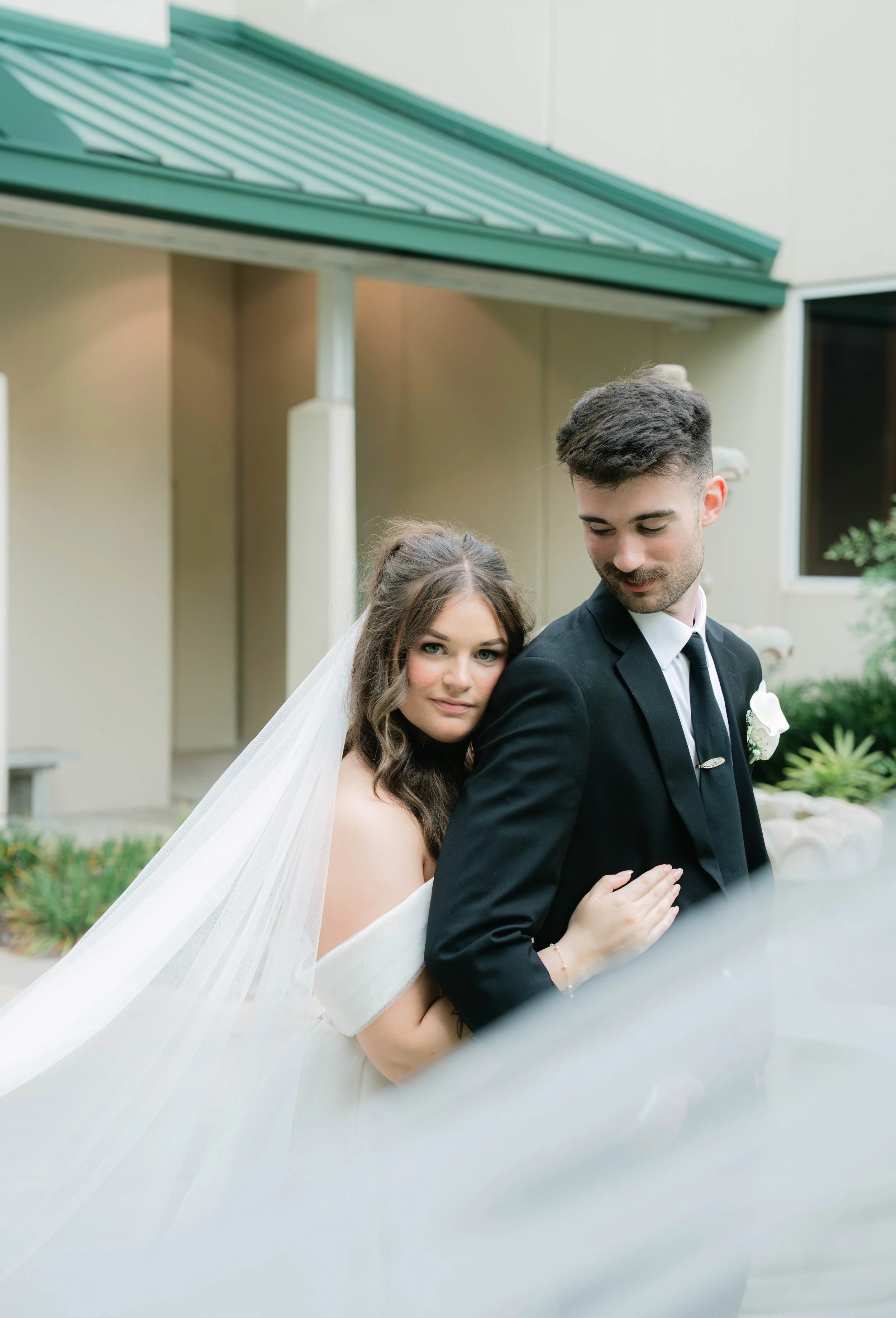 A newlywed couple posing outdoors, the bride with a long veil and the groom in a black suit, standing close together with the bride resting her hand on the groom's arm.