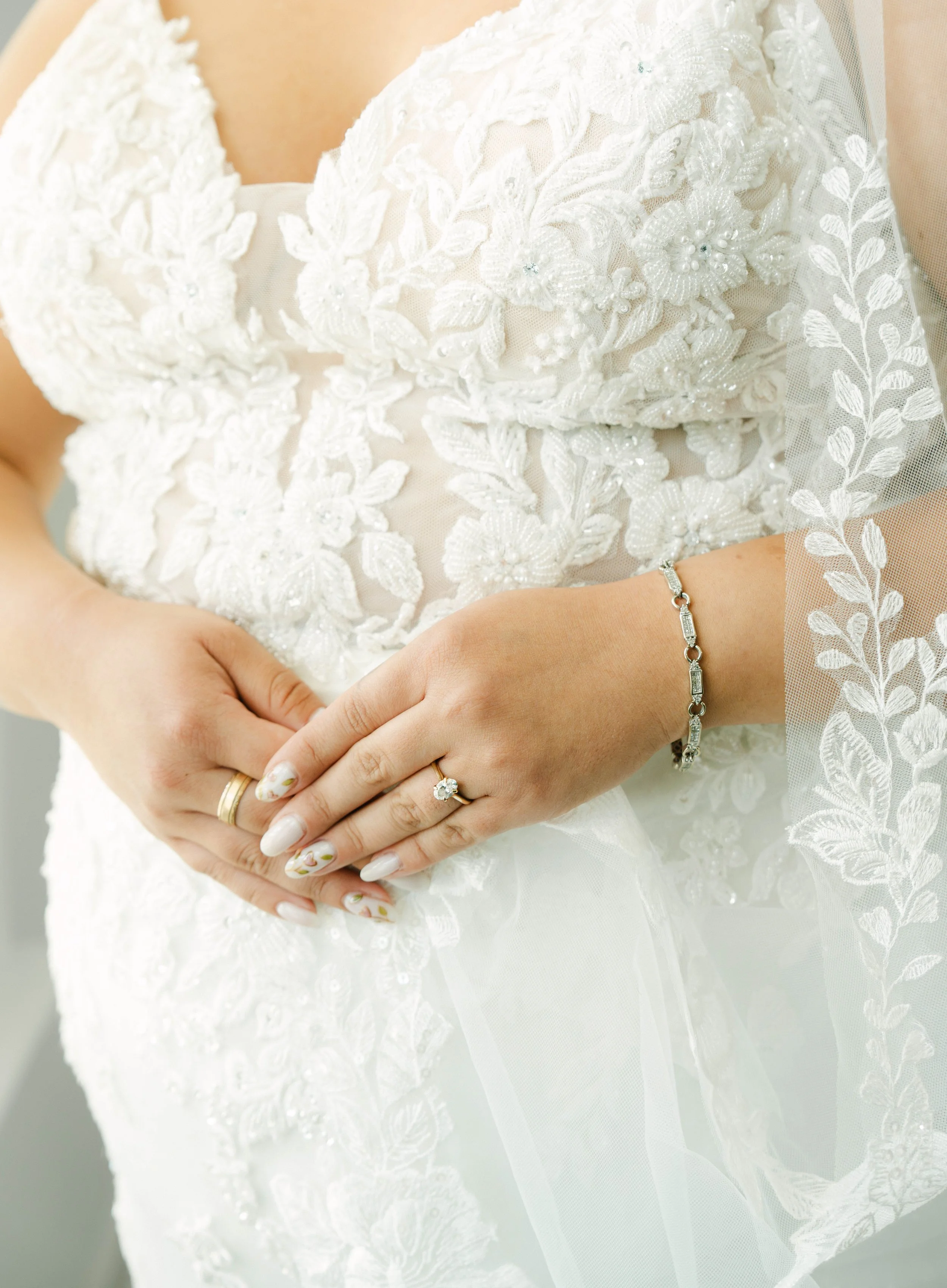 Close-up of a bride's hands resting on her waist, wearing a white lace wedding dress with floral embroidery and pearl embellishments. She is also wearing rings and a delicate bracelet.