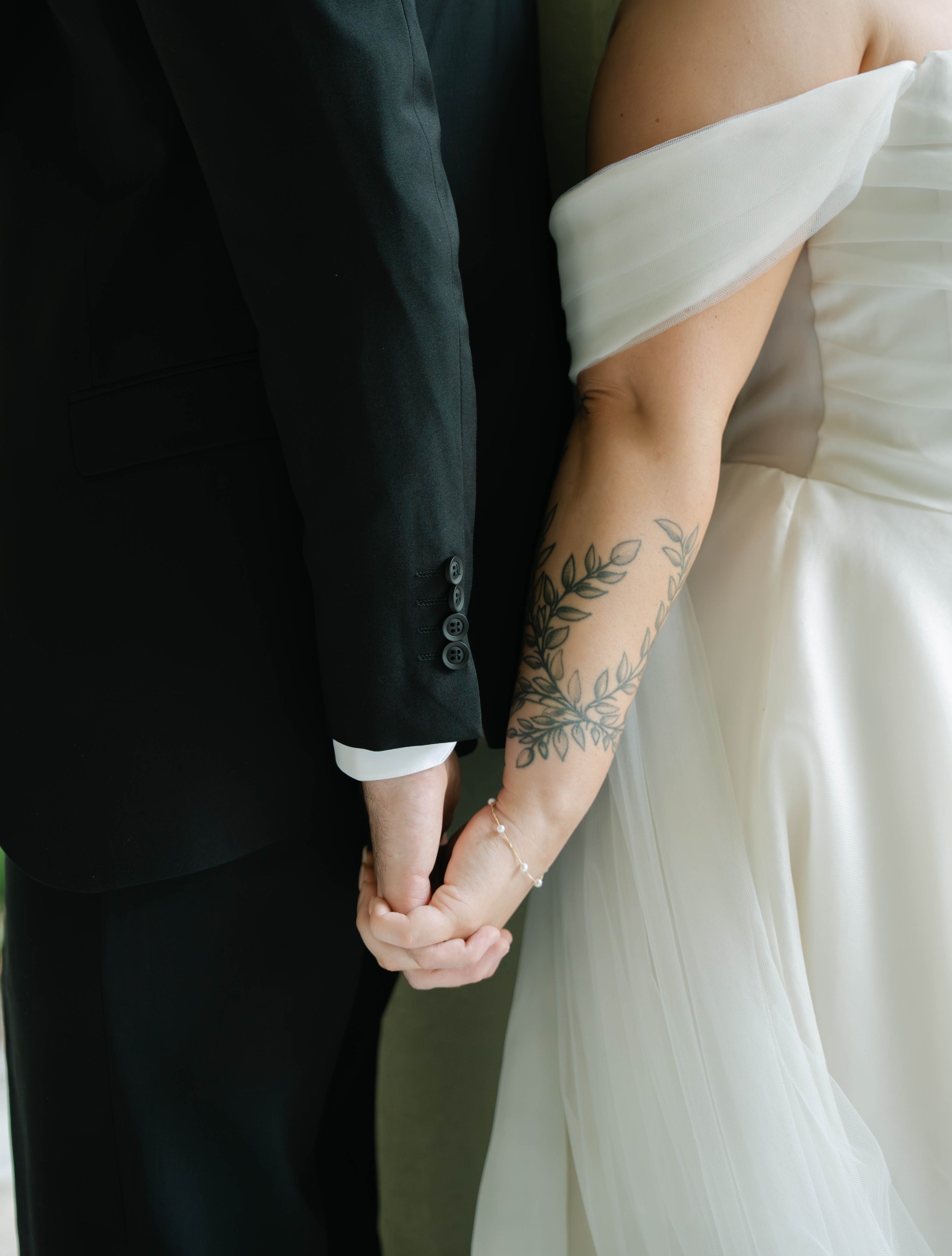 A couple dressed in formal attire holding hands, with the woman wearing a white dress with a tattoo on her arm and a bracelet, and the man wearing a black suit.