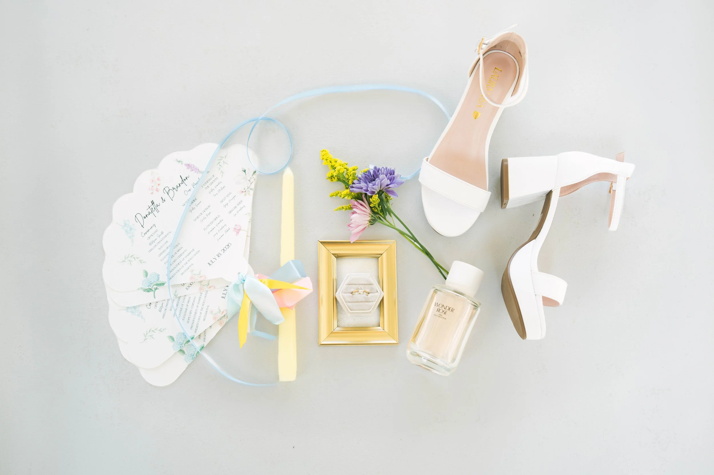 Wedding accessories including white high-heeled shoes, a wedding invitation, a bouquet of flowers, a gold picture frame, a perfume bottle, and an engagement ring in a box, arranged on a white surface.