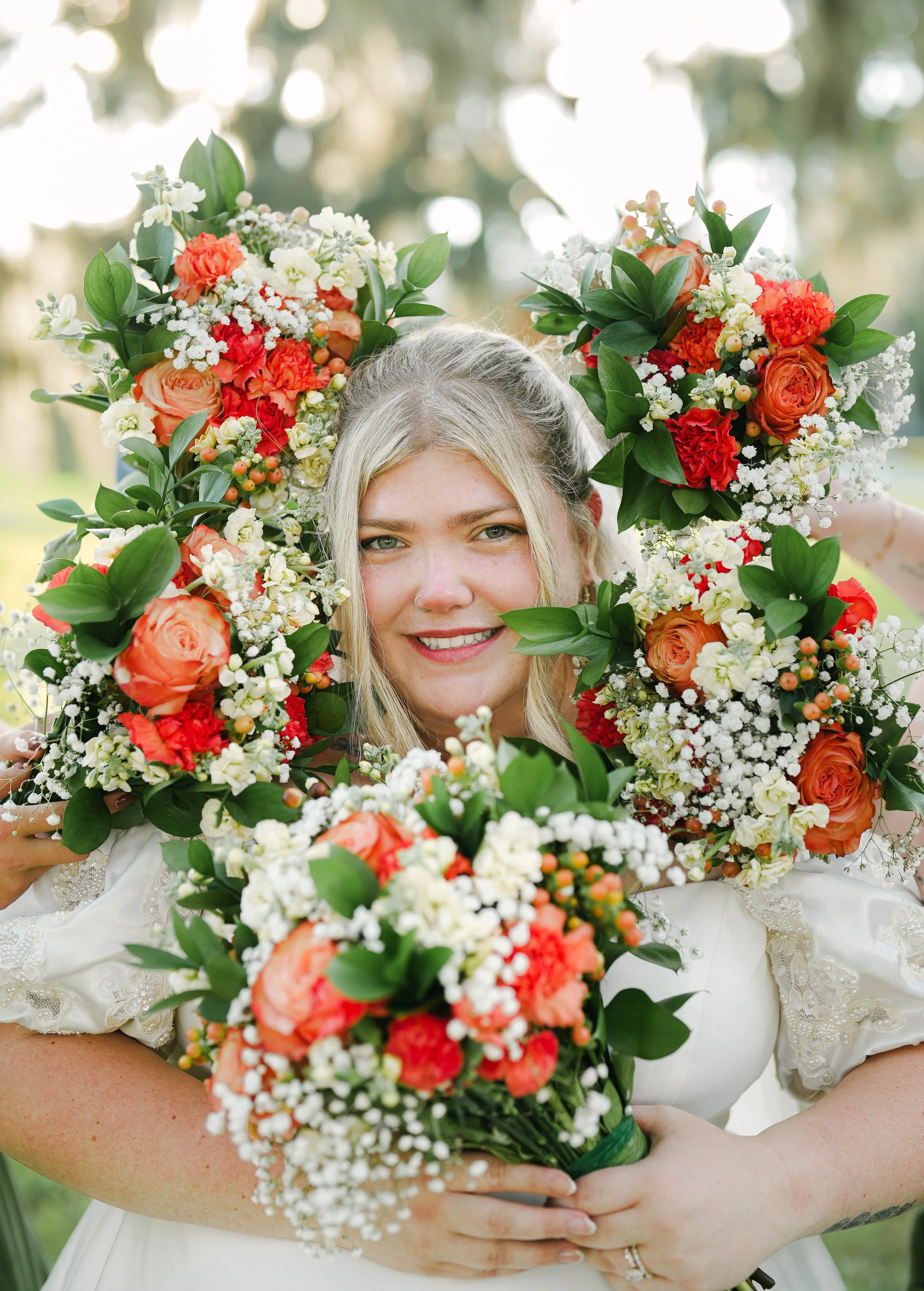 A woman with gray hair and a nose ring holds a bouquet of red, orange, and white flowers, with greenery, in front of her face outdoors.