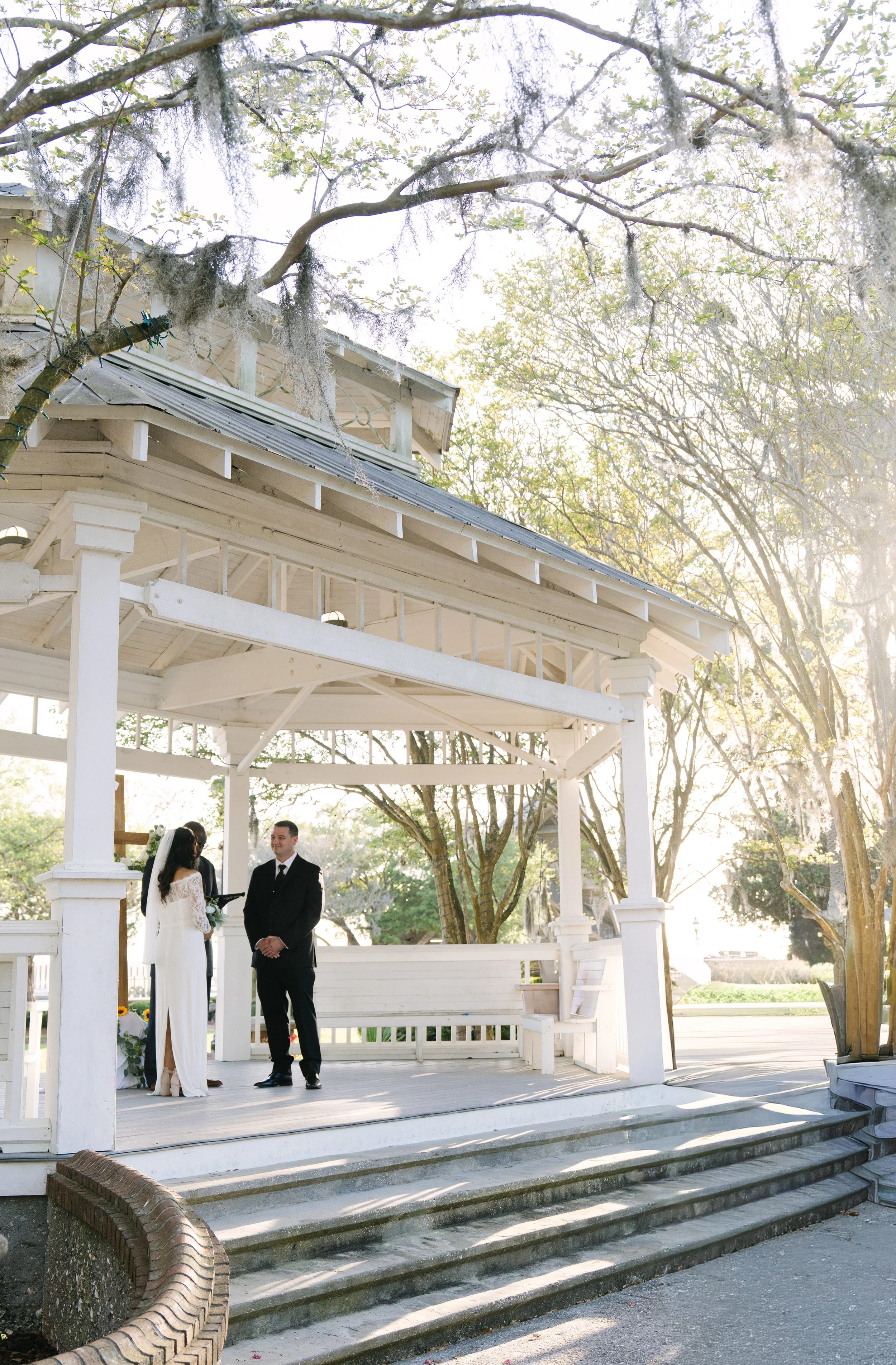 A bride and groom standing in front of an officiant during a wedding ceremony outdoors, under a white gazebo with trees and sunlight in the background.