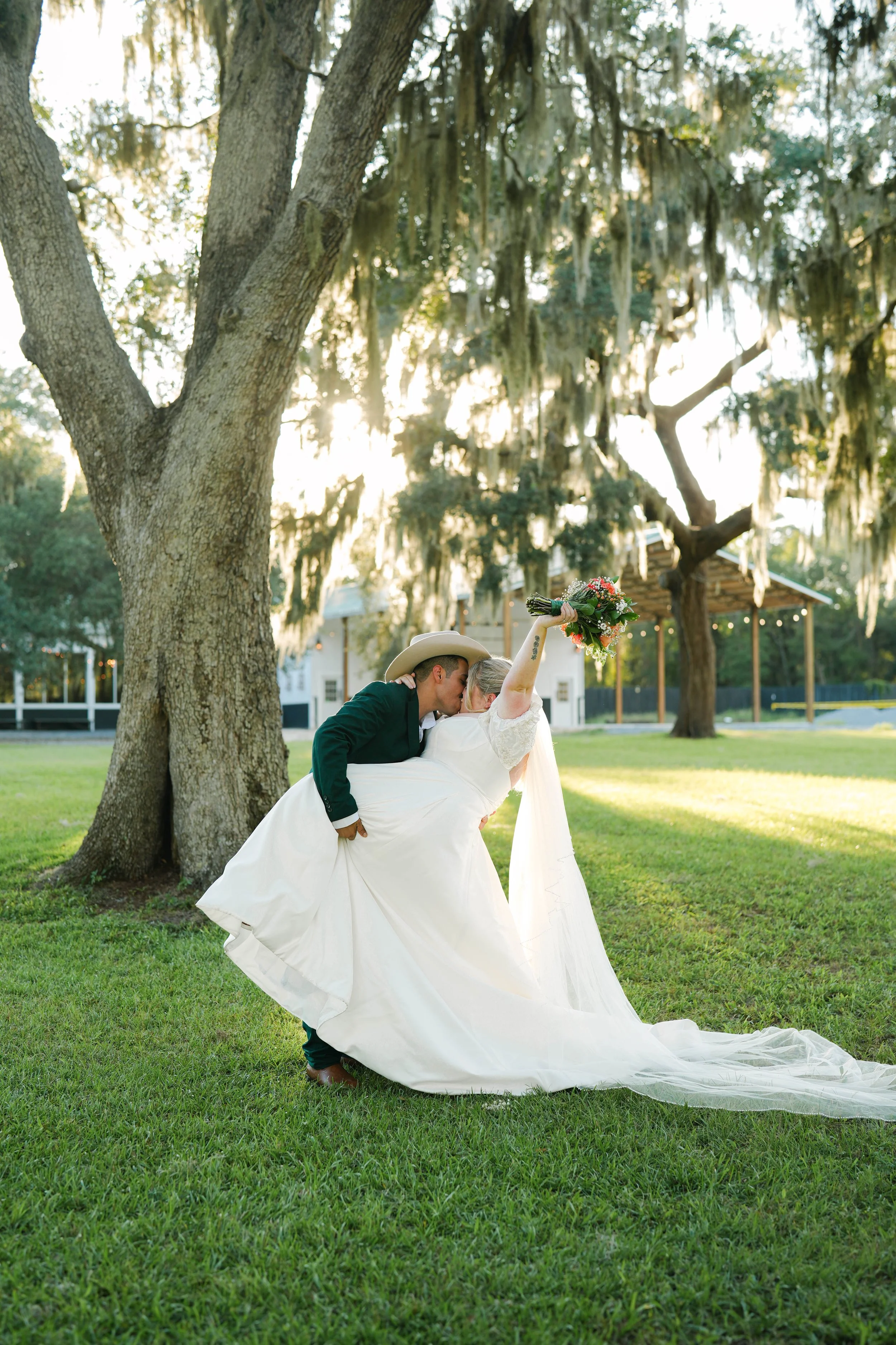 A groom and bride dancing outdoors on grass, with the groom lifting the bride and kissing her cheek. The bride is holding a bouquet, and the scene is illuminated by sunlight with trees and a building in the background.