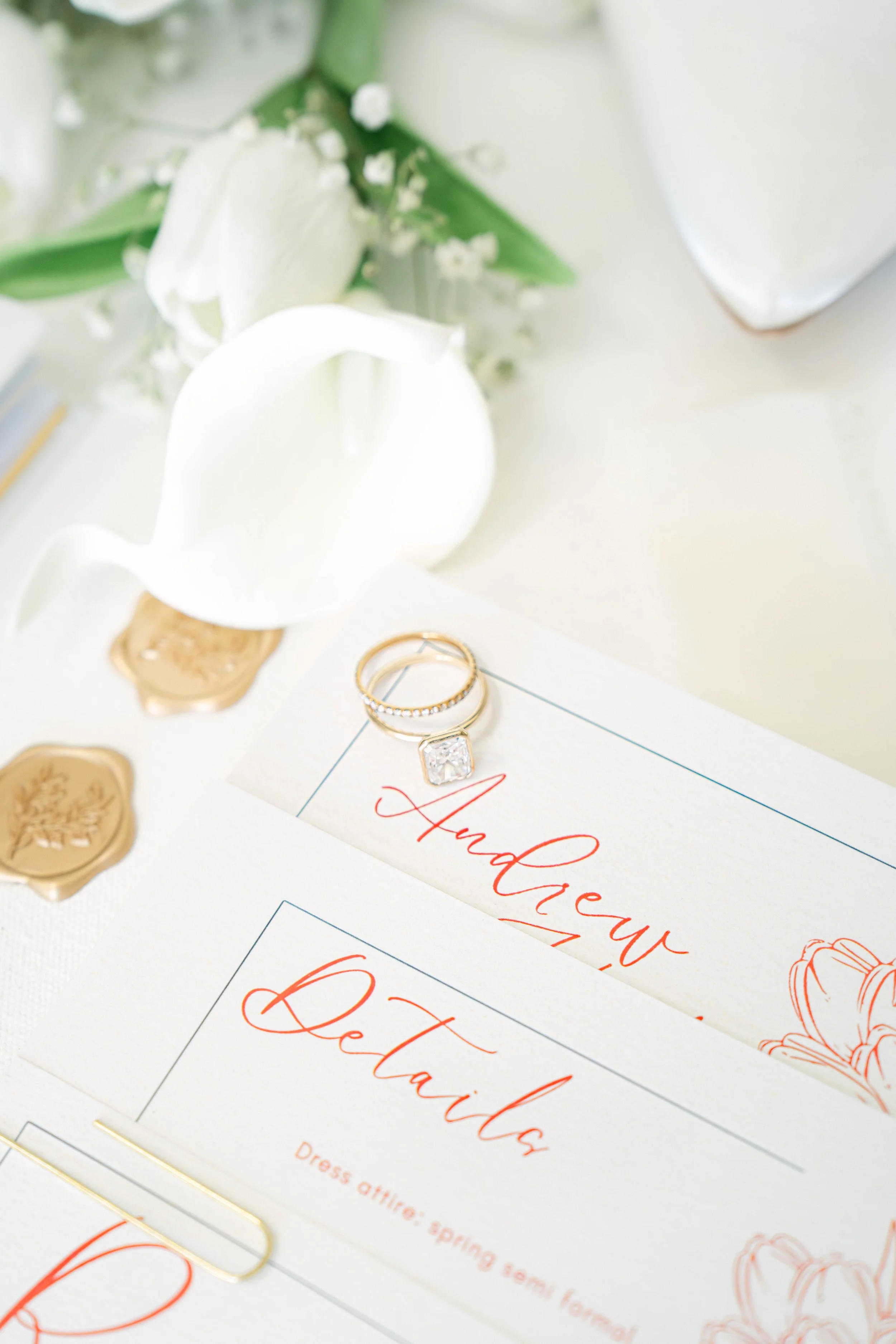 Elegant white calla lily flower flower with green leaves, surrounding wedding rings, gold seals, and a wedding invitation with red cursive text.